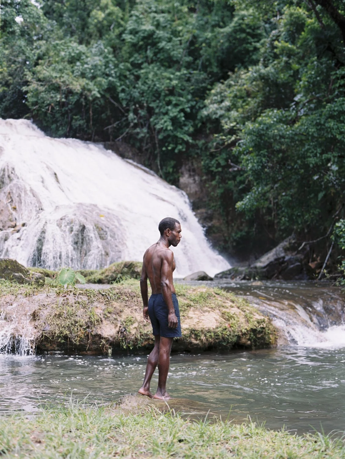 Man standing on a rock in a river near a waterfall surrounded by lush green trees.