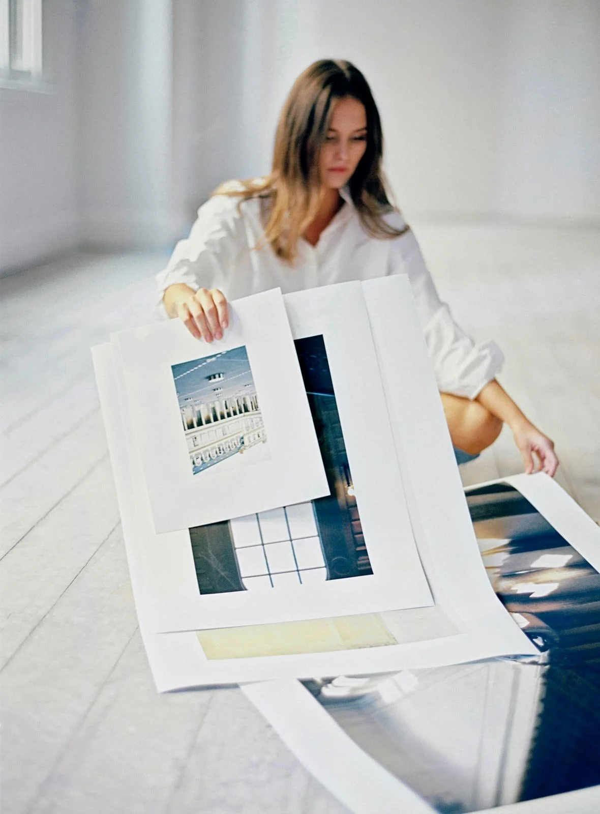 A woman sitting on the floor looking at photographs and prints laid out in front of her, with some held in her hand.