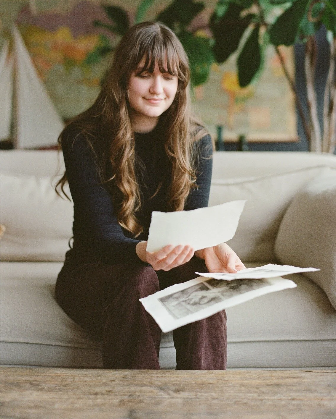 A young woman with long brown hair and bangs, dressed in a black top and maroon pants, sitting on a light-colored sofa, looking at black-and-white photographs and papers in her hands. She is in a cozy room with a wooden floor, large green plants, and