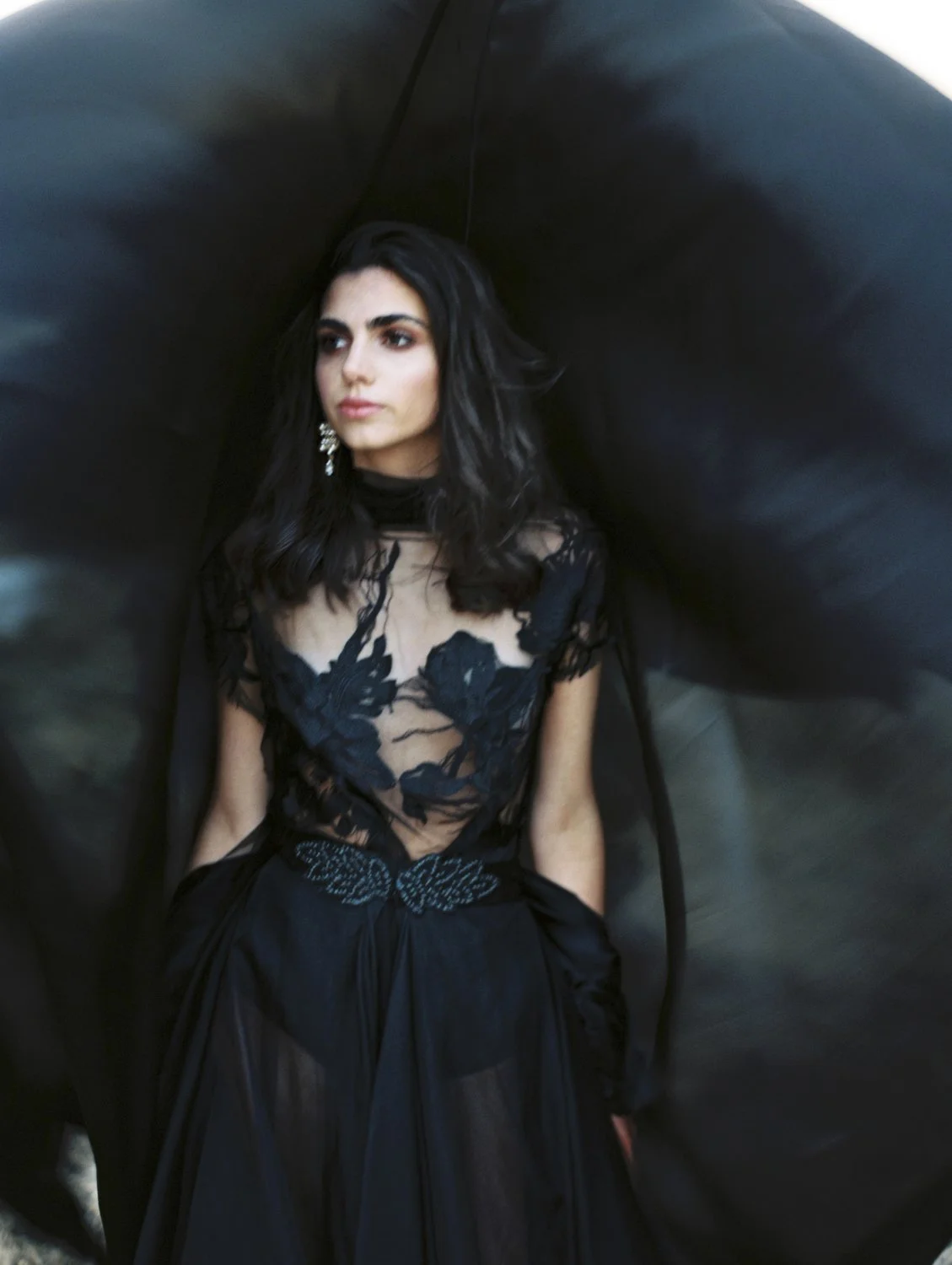 A woman with dark hair wearing a black lace dress with floral embroidery, standing with her hands in her pockets and looking to the side, with a dark, flowing fabric backdrop behind her.