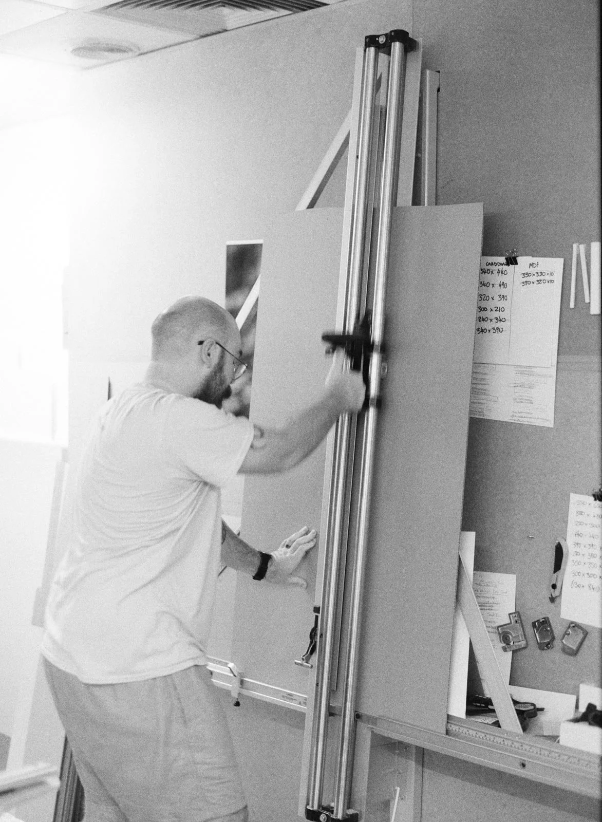 A man with glasses and a short beard in a white shirt and shorts is operating a large sliding table saw in a woodworking workshop, cutting a piece of material.