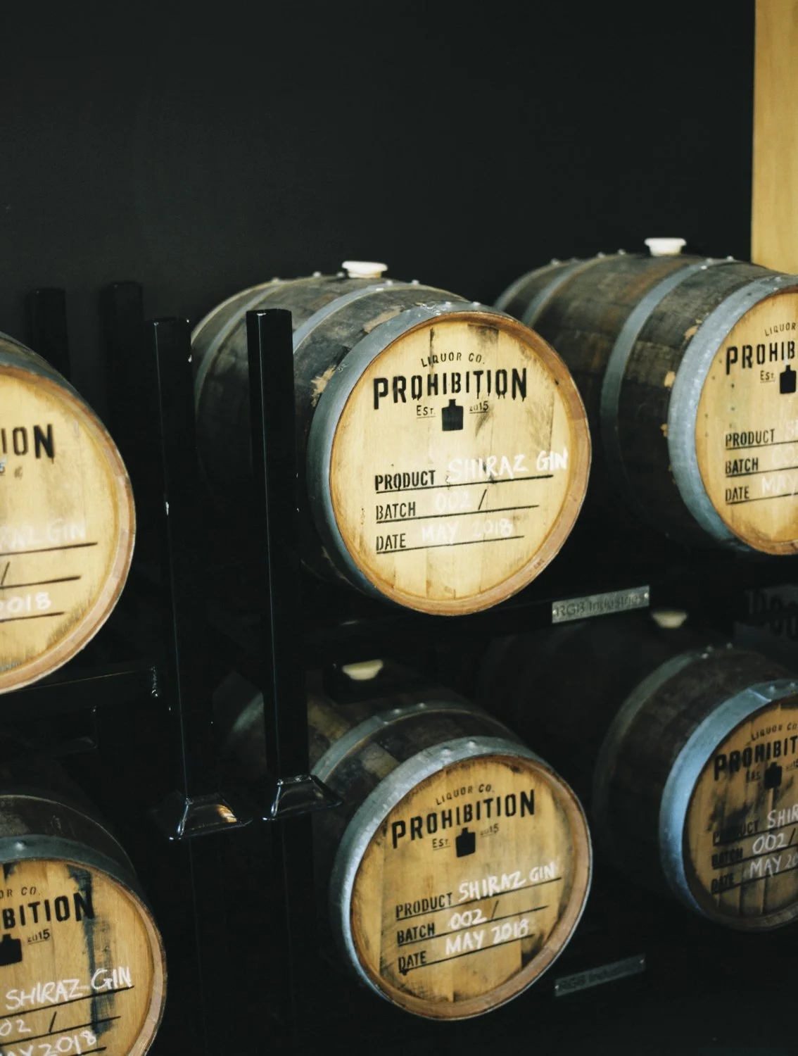 Wooden barrels labeled 'Prohibition Liquor Co.' with handwritten labels indicating 'Shiraz Gin,' batch number 002, and date May 2018, stored on black metal racks.