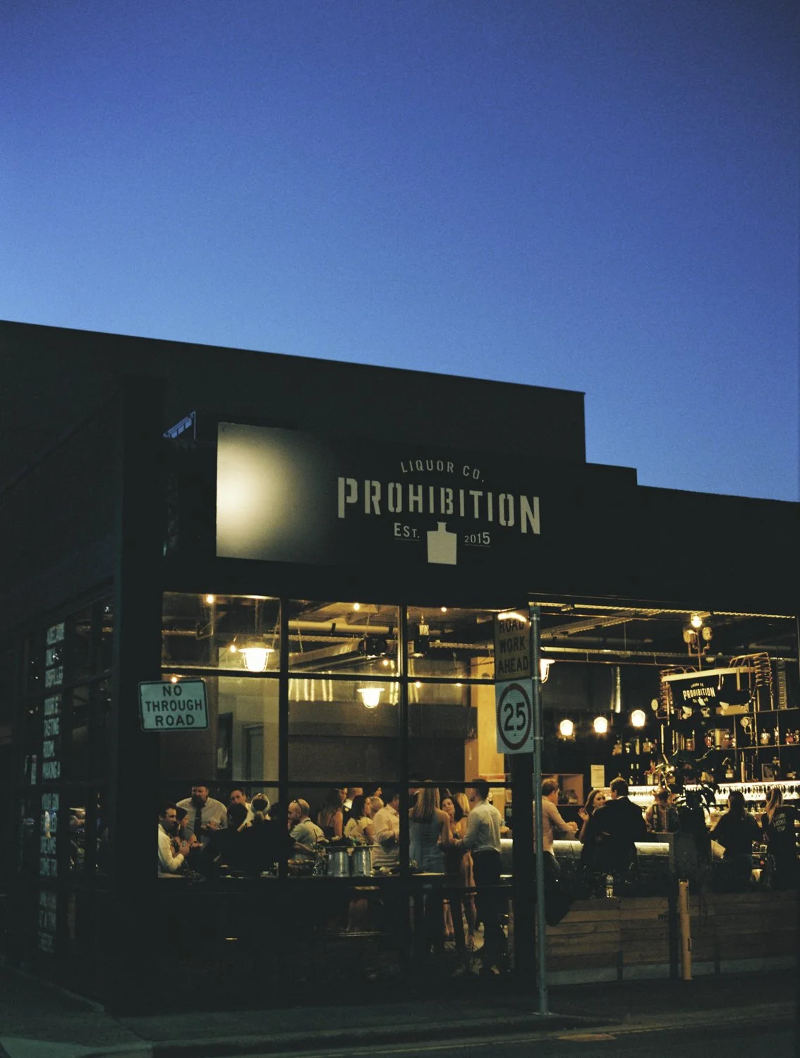 The exterior of a bar or restaurant called Prohibition, established in 2015, with people inside, nighttime sky, and street signs visible.