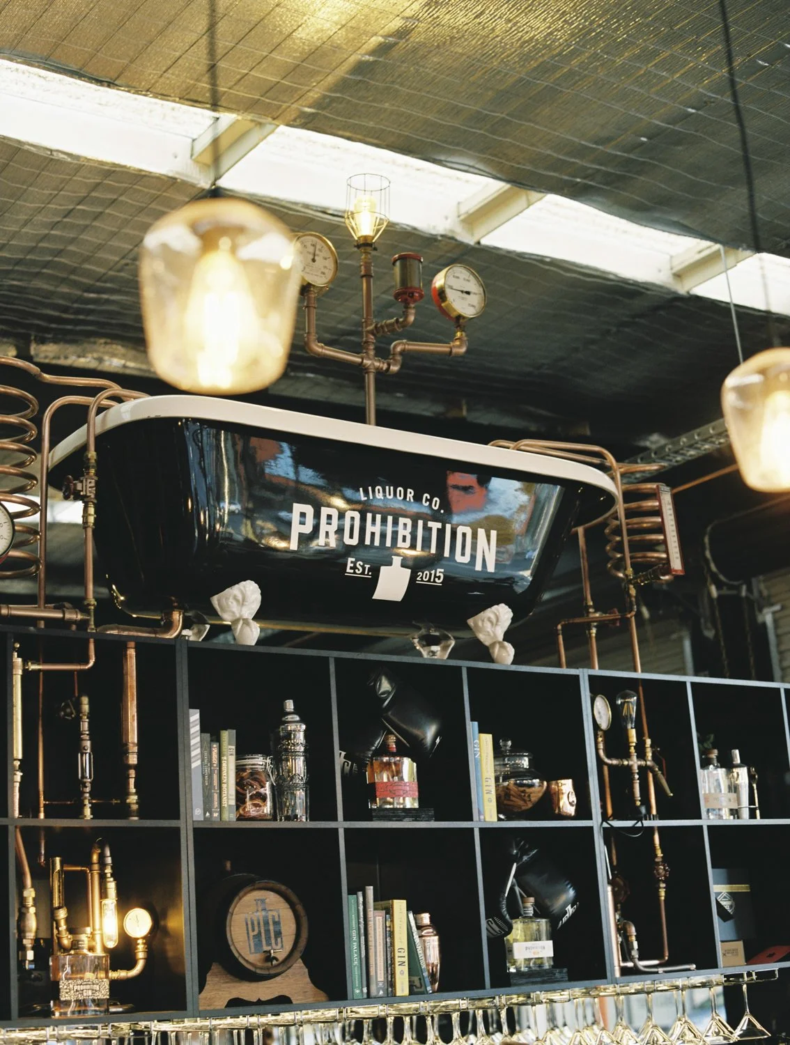 Interior of a bar with steampunk decor, including copper pipes, gauges, a black bathtub labeled 'Prohibition,' books, and bottles on shelves.
