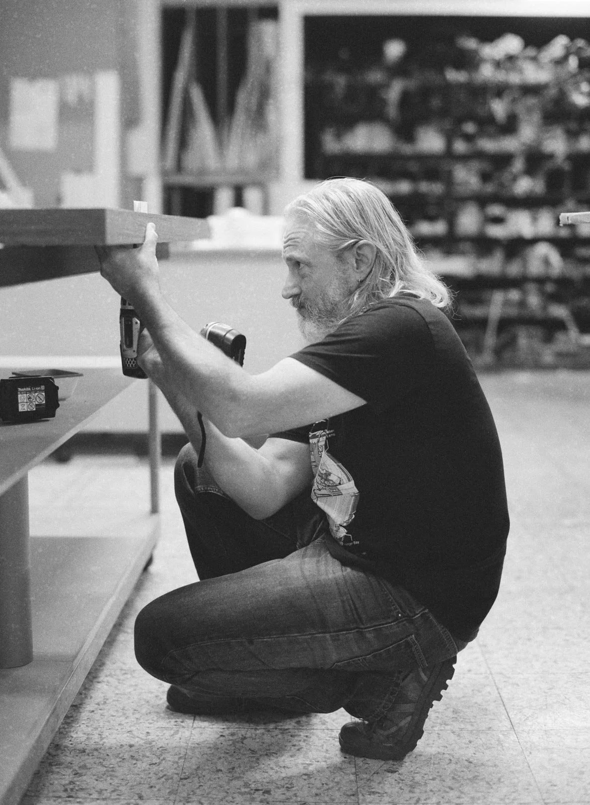 A man with long hair and a beard kneeling on the floor, holding a power drill, working on a wooden shelf in a workshop.