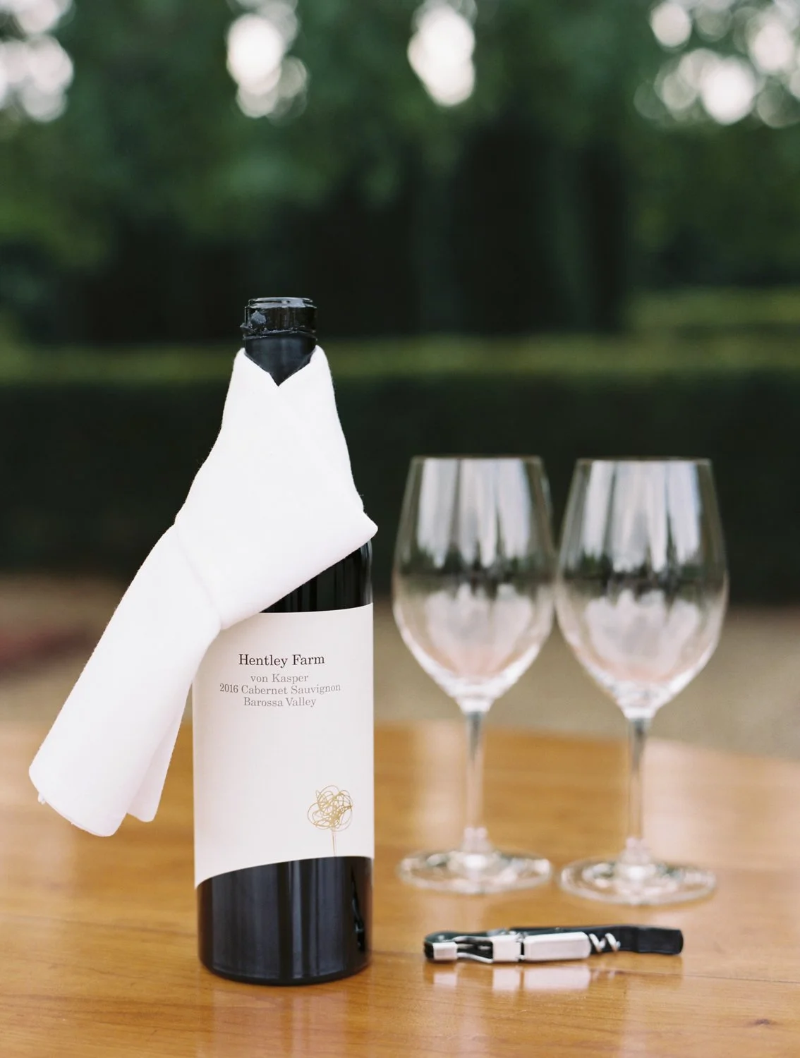 A bottle of wine with a white cloth wrapped around its neck, two empty wine glasses, and a corkscrew on a wooden table outdoors with a blurred green background.