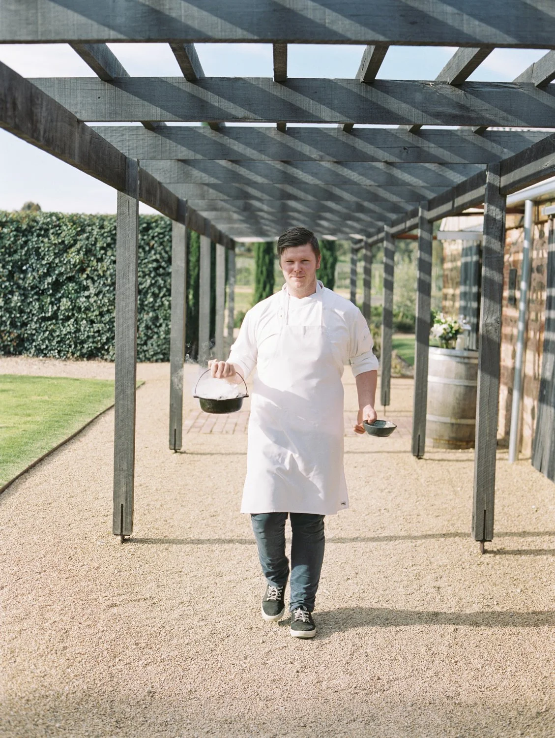 Male chef walking outside under a wooden pergola, wearing a white apron and holding a small cast iron skillet and a bowl.
