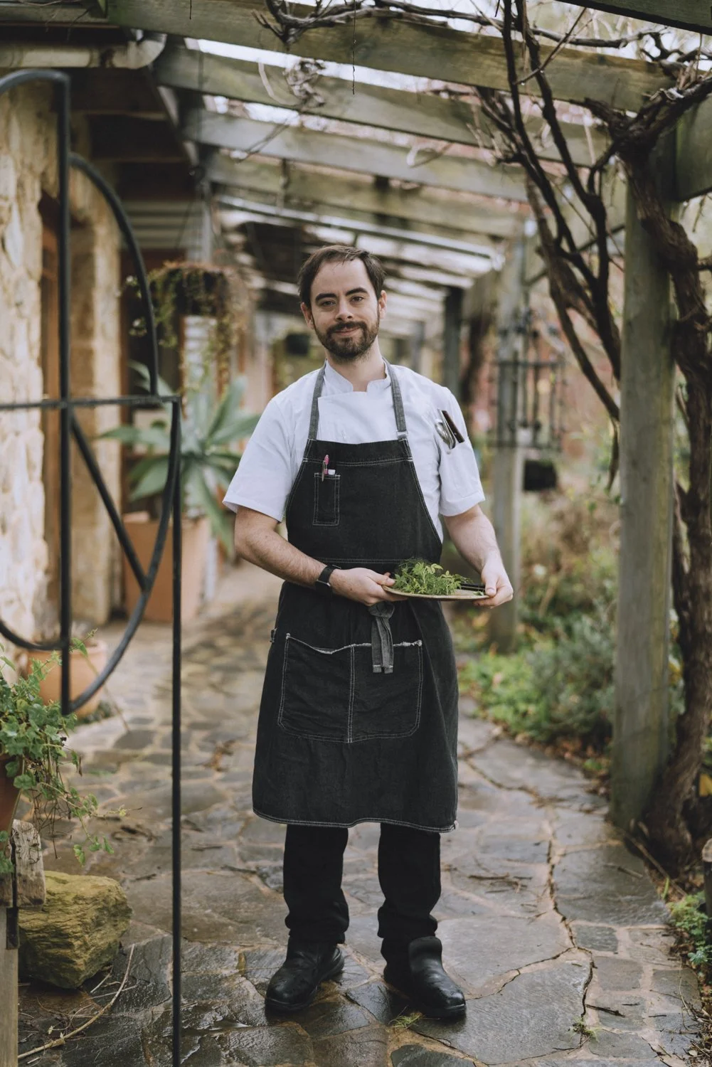 Male chef standing outdoors on a stone pathway, holding a tray of fresh herbs, under a wooden pergola with plants and vines.