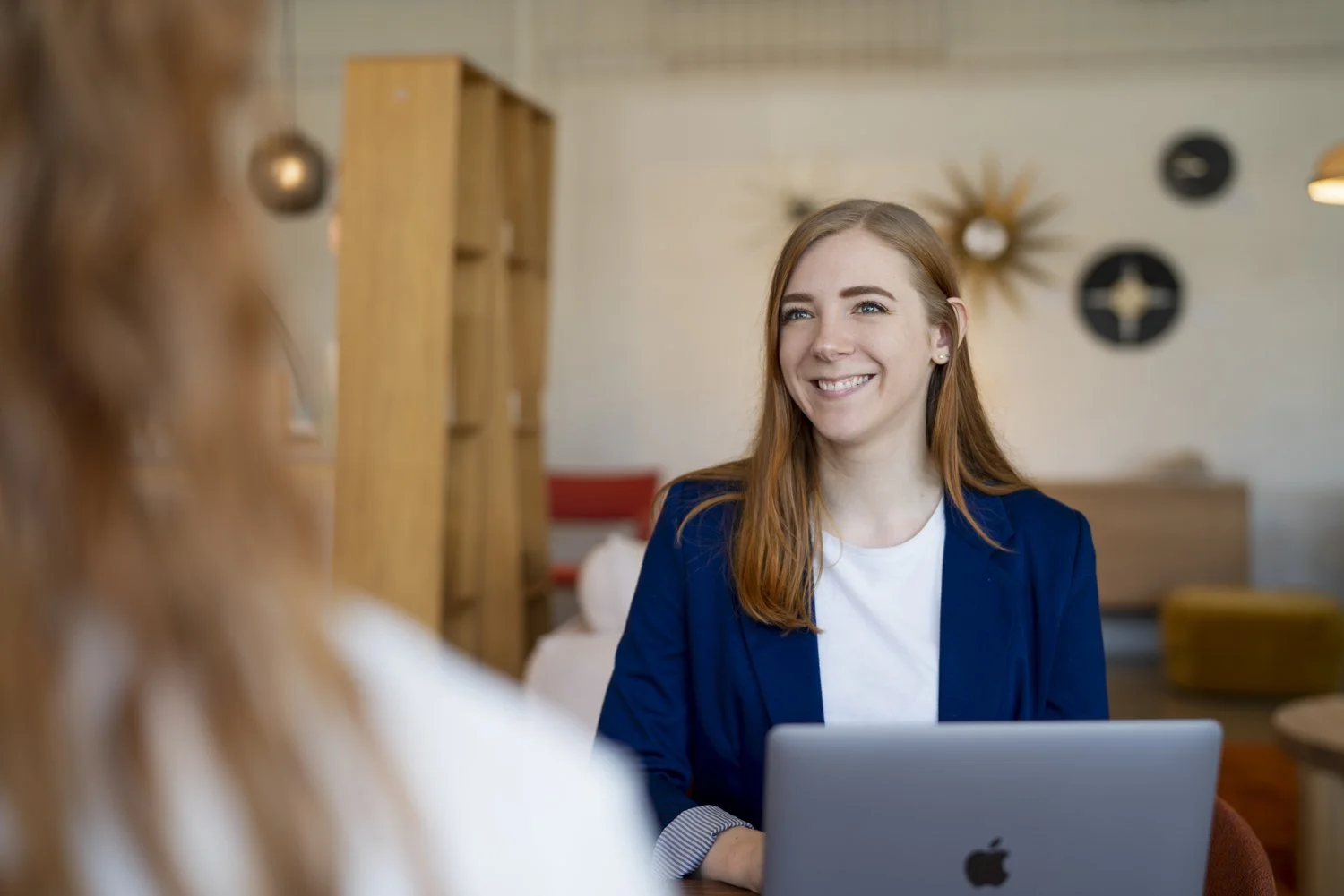 A woman with long red hair smiling in a meeting with another person visible from behind, sitting at a table with a laptop in a modern, decorated space.
