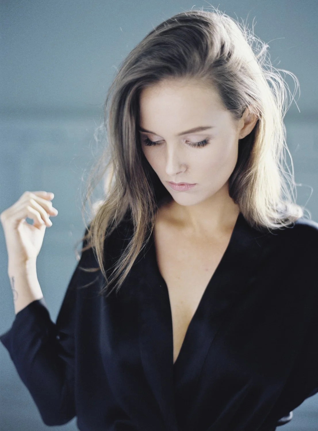 A woman with long, light brown hair looking down, wearing a black top with a deep V neckline against a soft blue background.