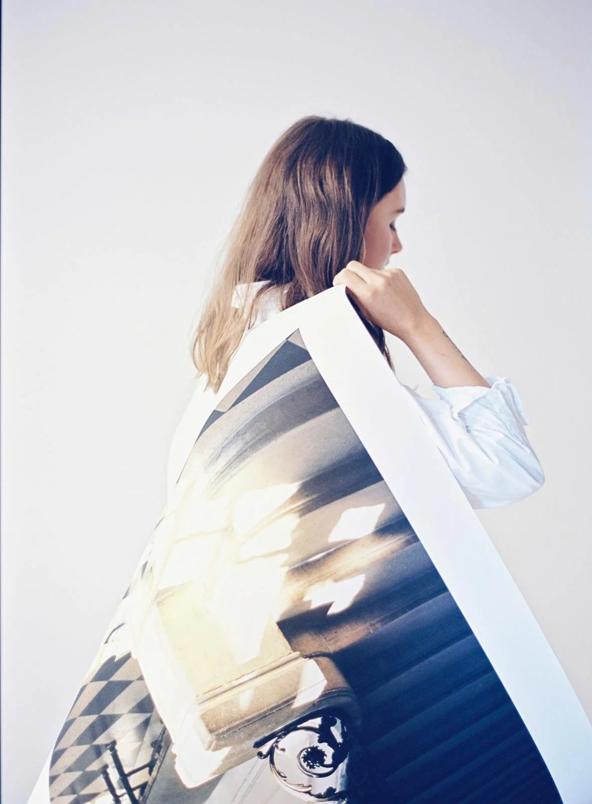 A woman with shoulder-length brown hair, wearing a white shirt, holding an oversized photograph of a historic building with arches and a street lamp, against a plain white background.
