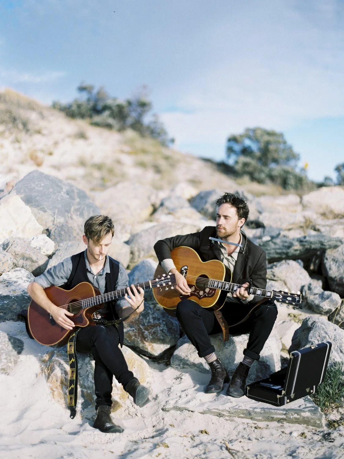 Two men playing guitars outdoors on a rocky beach, with a blue sky and distant trees in the background.