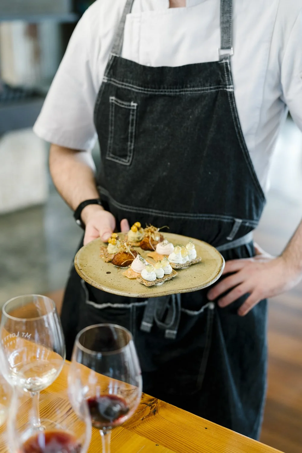 Person wearing a white shirt and black apron holding a plate of assorted small appetizers or canapés in a restaurant setting.