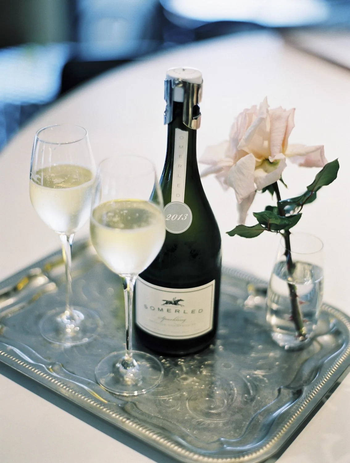 A bottle of Somerled sparkling wine on a silver tray with two glasses filled with champagne, a single pink rose in a small vase, and a white tablecloth in the background.