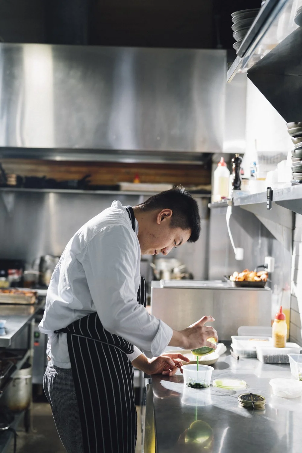Chef preparing food in a commercial kitchen, pouring sauce into a container on a stainless steel countertop.