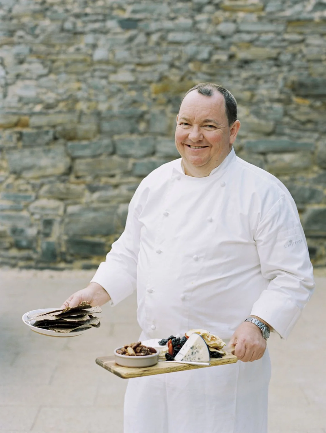 A smiling chef in a white uniform holding a wooden tray with cheese, nuts, berries, and crackers, standing in front of a stone wall.