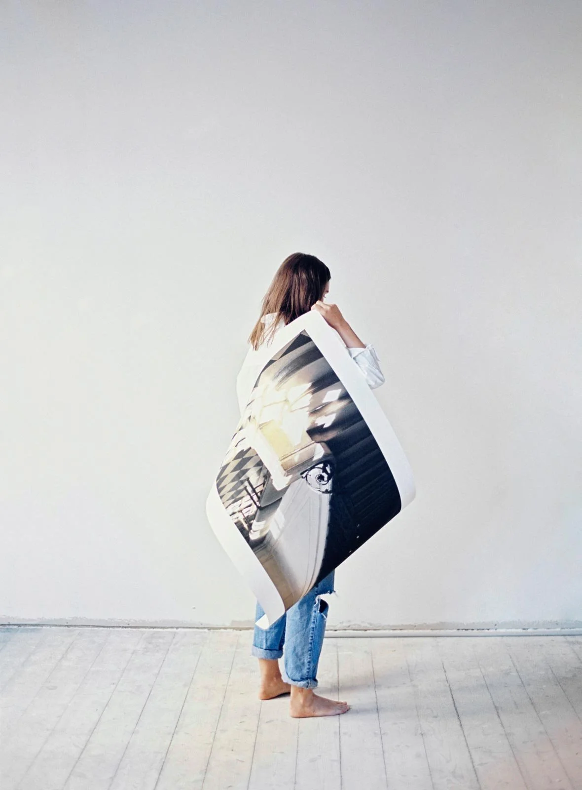 A person with long hair wearing jeans is holding a large, distorted photograph in front of their body, standing barefoot against a plain white wall and wooden floor.