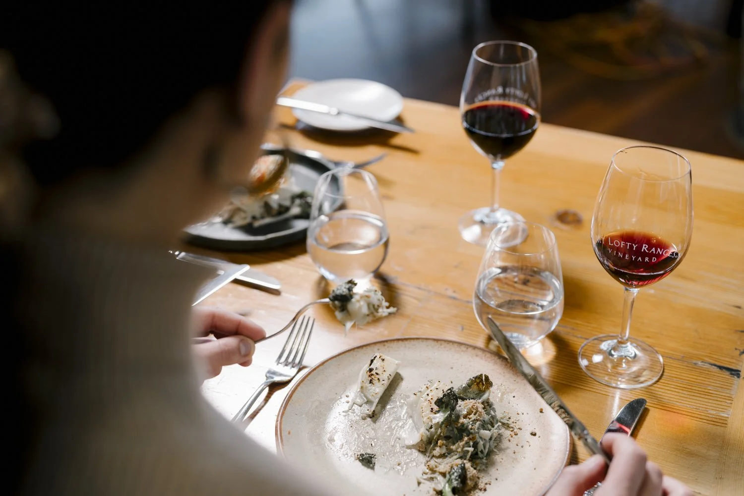 Person eating at a wooden table with partially eaten plate, two glasses of red wine, and two glasses of water, in a cozy setting.