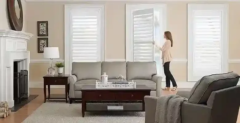 Living room with a beige sofa, armchair, coffee table, table lamp, fireplace, and large white shutters. A woman adjusts the shutters near a window.