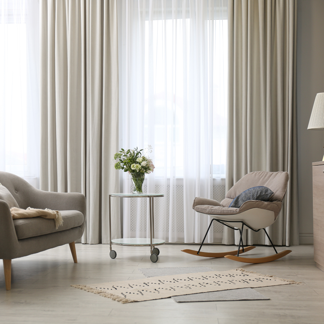 Modern living room with a gray sofa, rocking chair, glass coffee table, vase of flowers, and light curtains.