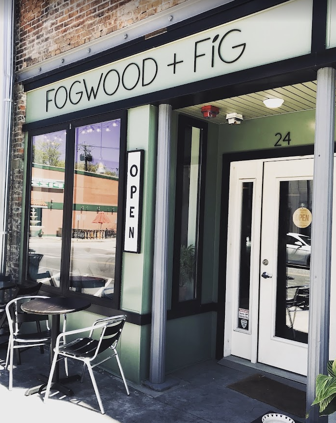 Exterior of a café named "Fogwood + Fig" with a large illuminated sign, an "Open" sign, a white door, and a small outdoor seating area with two metal chairs and a table.