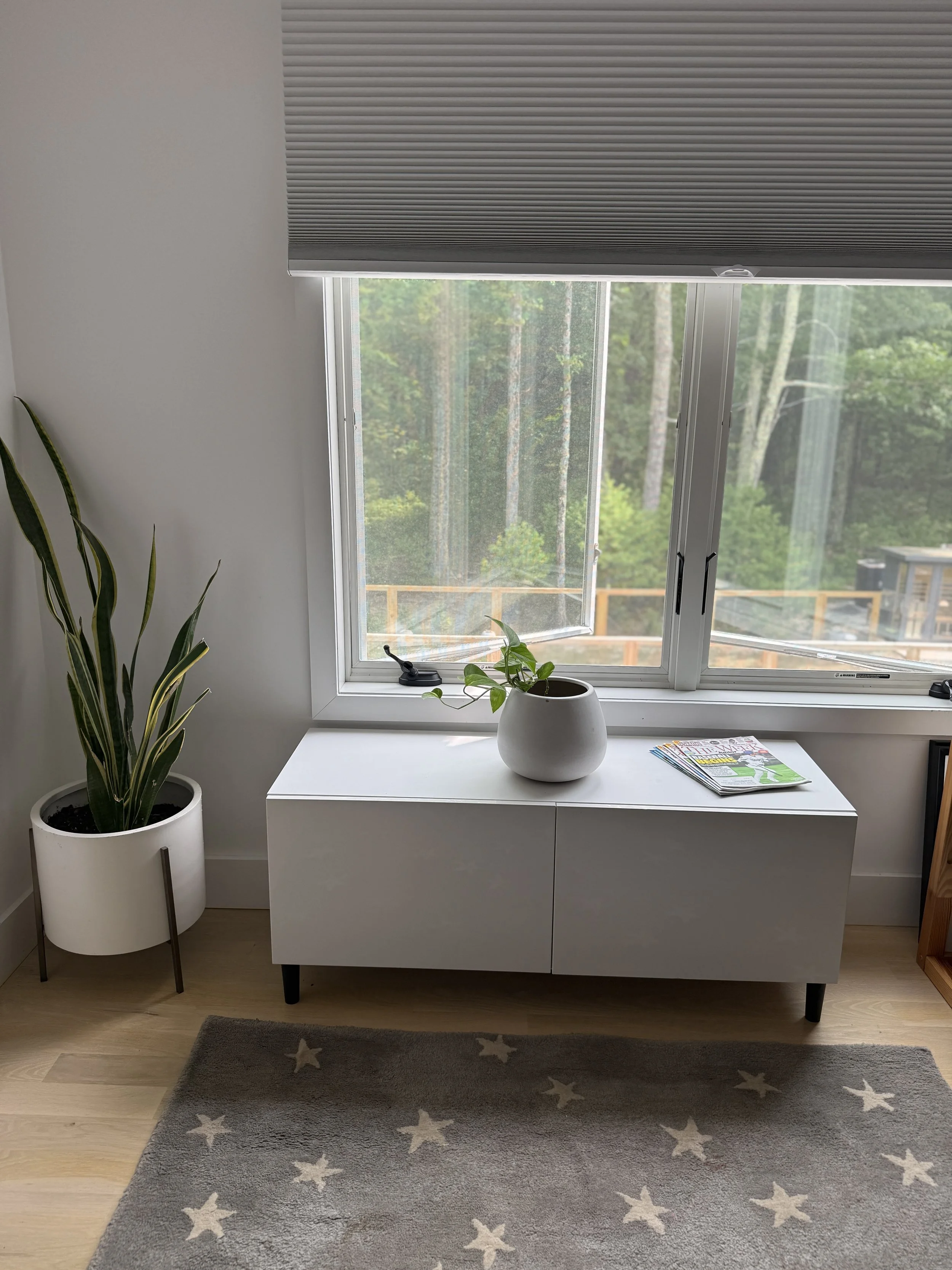 A cozy corner with a white cabinet beneath a large window, which has a partially open blind. On the cabinet, there is a round white pot with a green plant, and some magazines are placed beside it. A tall, striped potted plant is to the left, and a st
