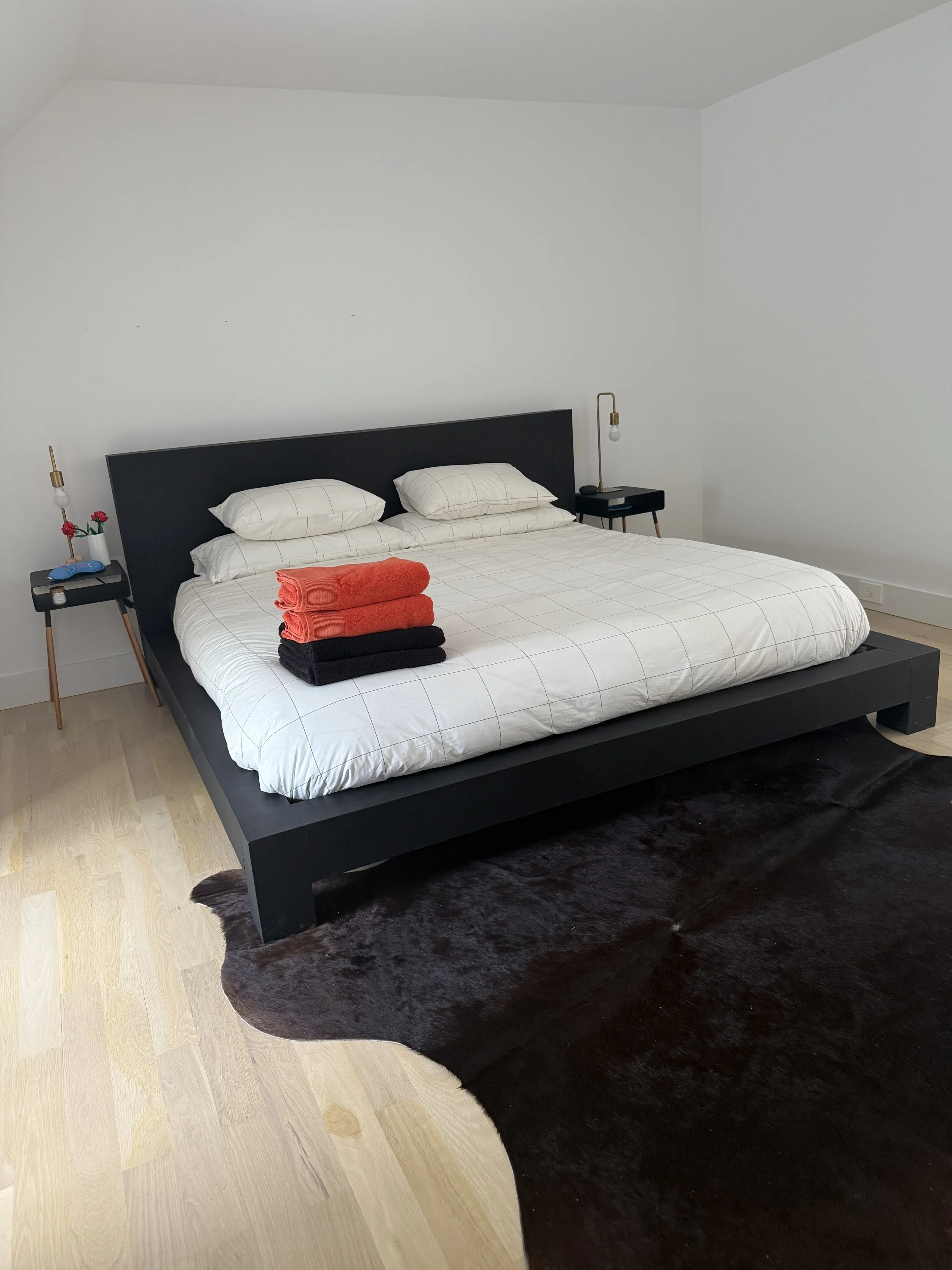 A modern bedroom with a black bed frame, white bedding, and four pillows. On the bed are three folded towels in red and black. There are two small black bedside tables with gold lamps on each side. The floor is light wood, and a dark cowhide rug is p