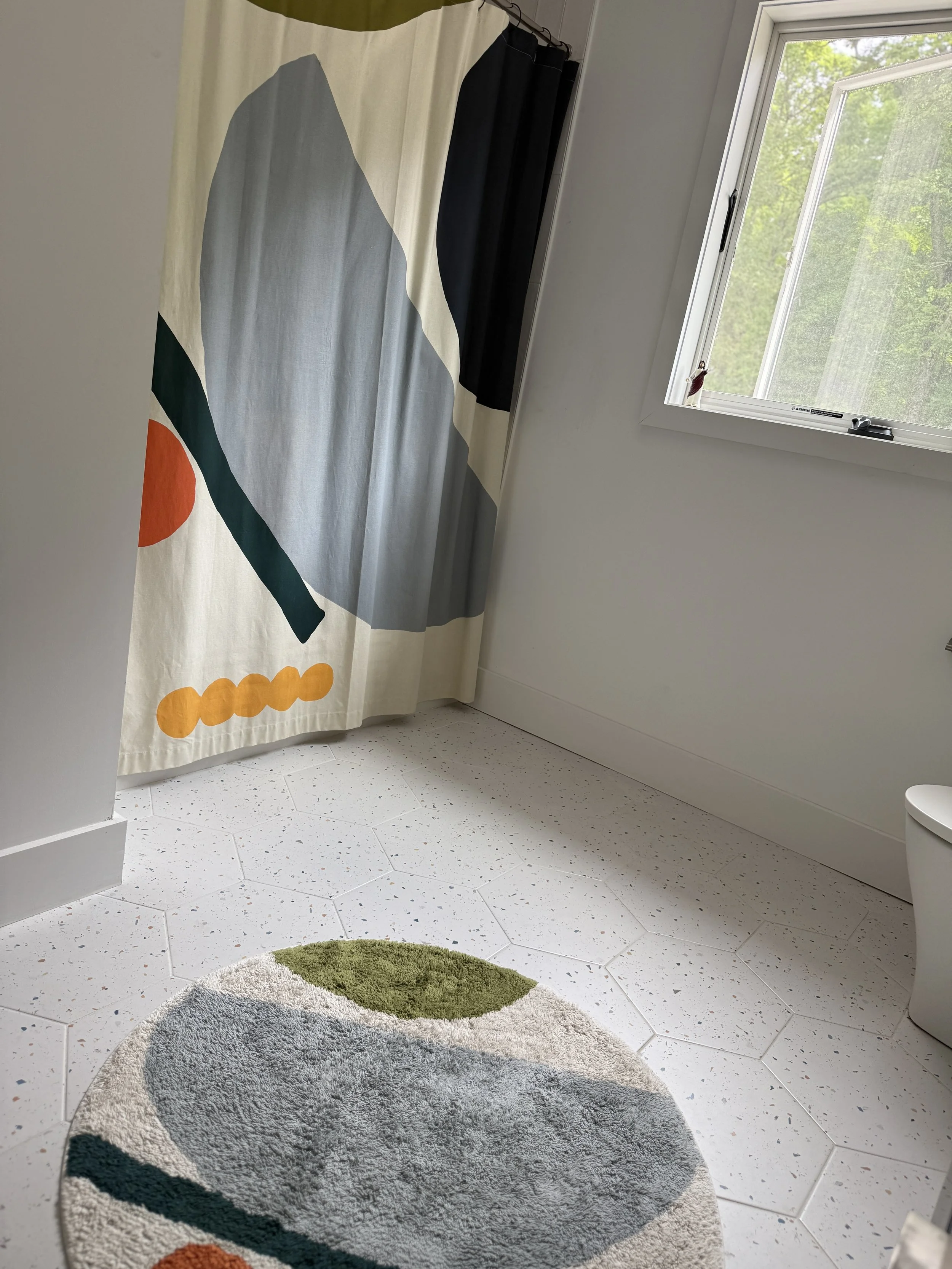 A bathroom with white hexagonal tile flooring, a round patterned rug, a window with natural light, and a shower curtain with abstract shapes in black, gray, orange, and green.