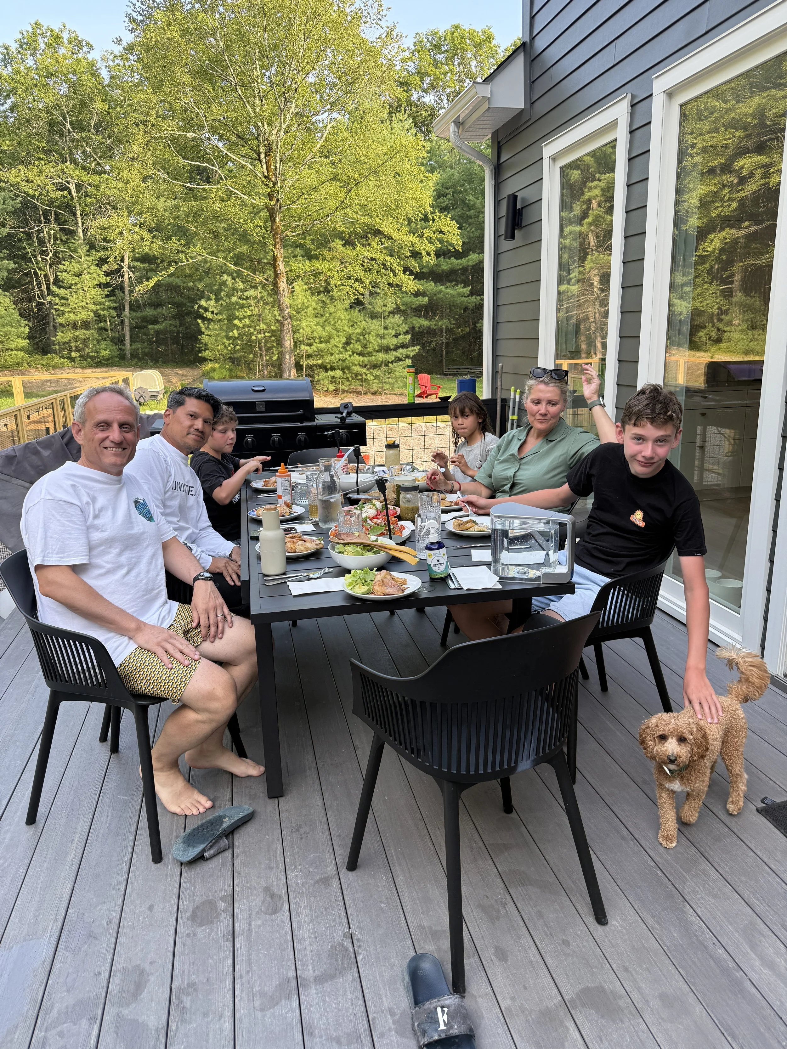 Family enjoying a backyard dinner on a deck with various food and drinks, with trees and a grill in the background, and a small dog standing near a young boy.