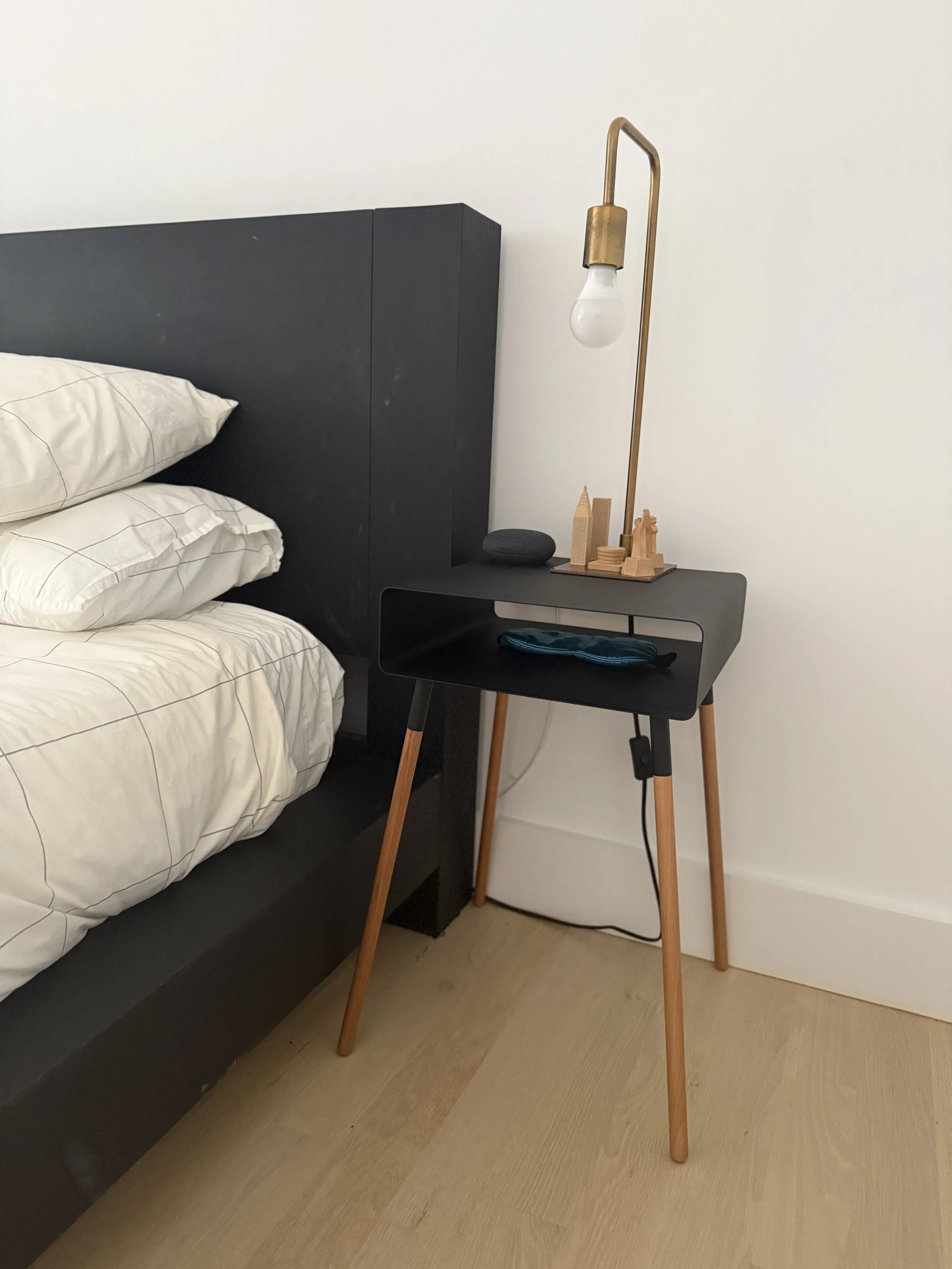 A black bedside table next to a bed with white bedding, featuring a small open shelf with a blue object, a modern black stone on top, and a decorative wooden cityscape. A brass floor lamp with a white bulb is placed on the table