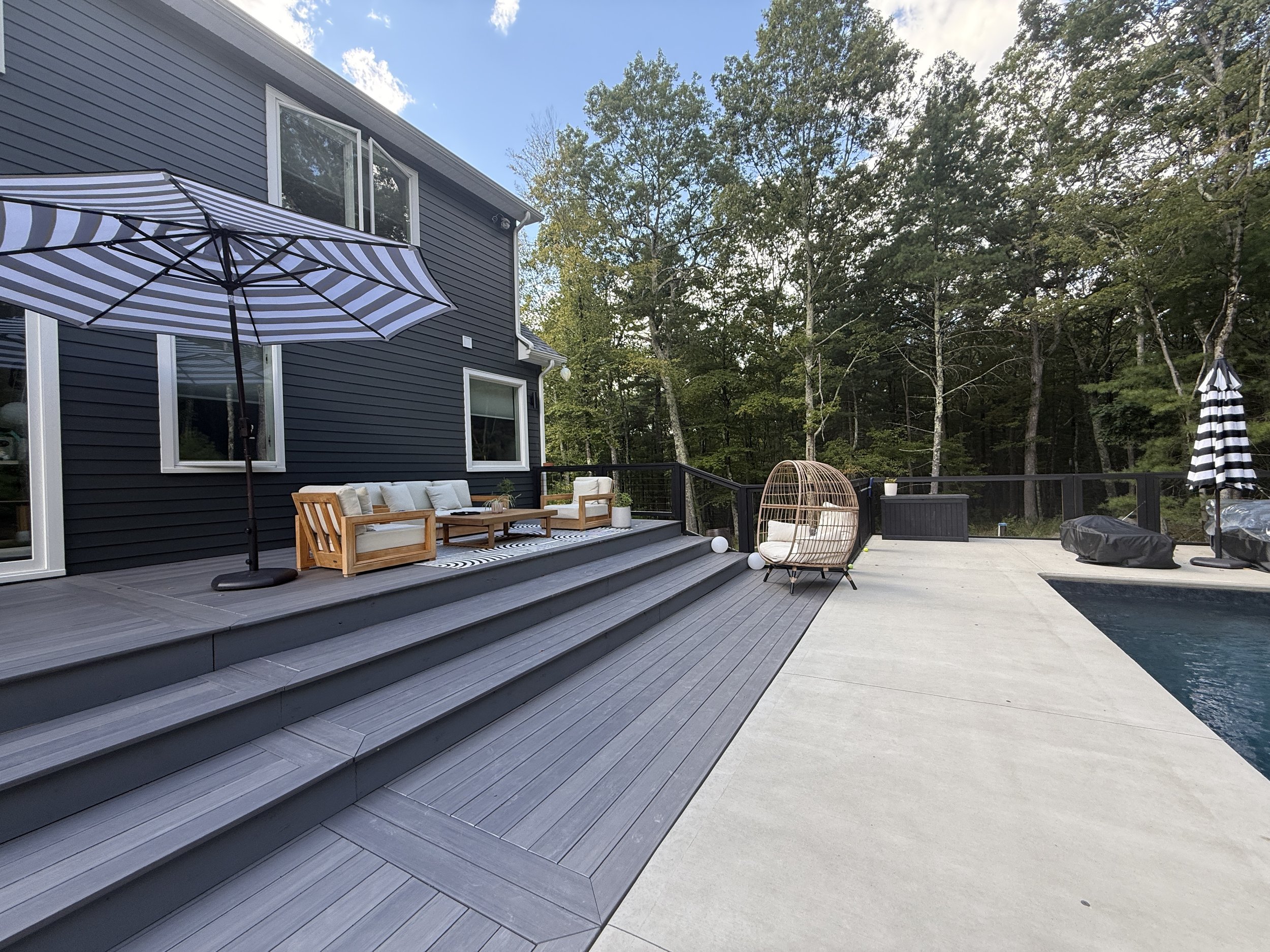 Outdoor patio area with wooden stairs, cushioned outdoor furniture, a rattan hanging chair, striped umbrellas, and a swimming pool, surrounded by trees.