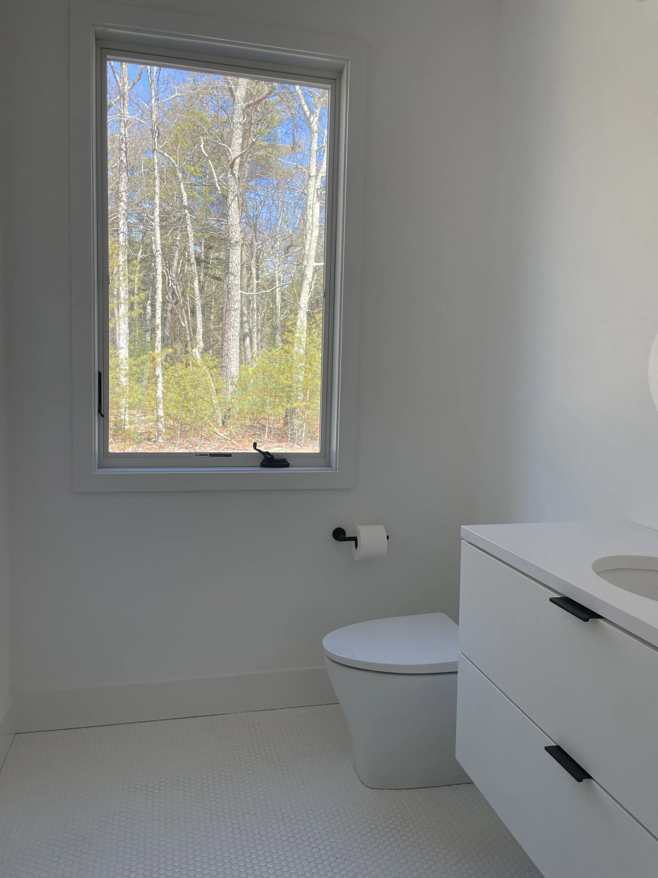 A modern, minimalist bathroom with a window showing a view of trees and a blue sky, a white toilet, a white vanity with black handles, and a wall-mounted toilet paper holder.