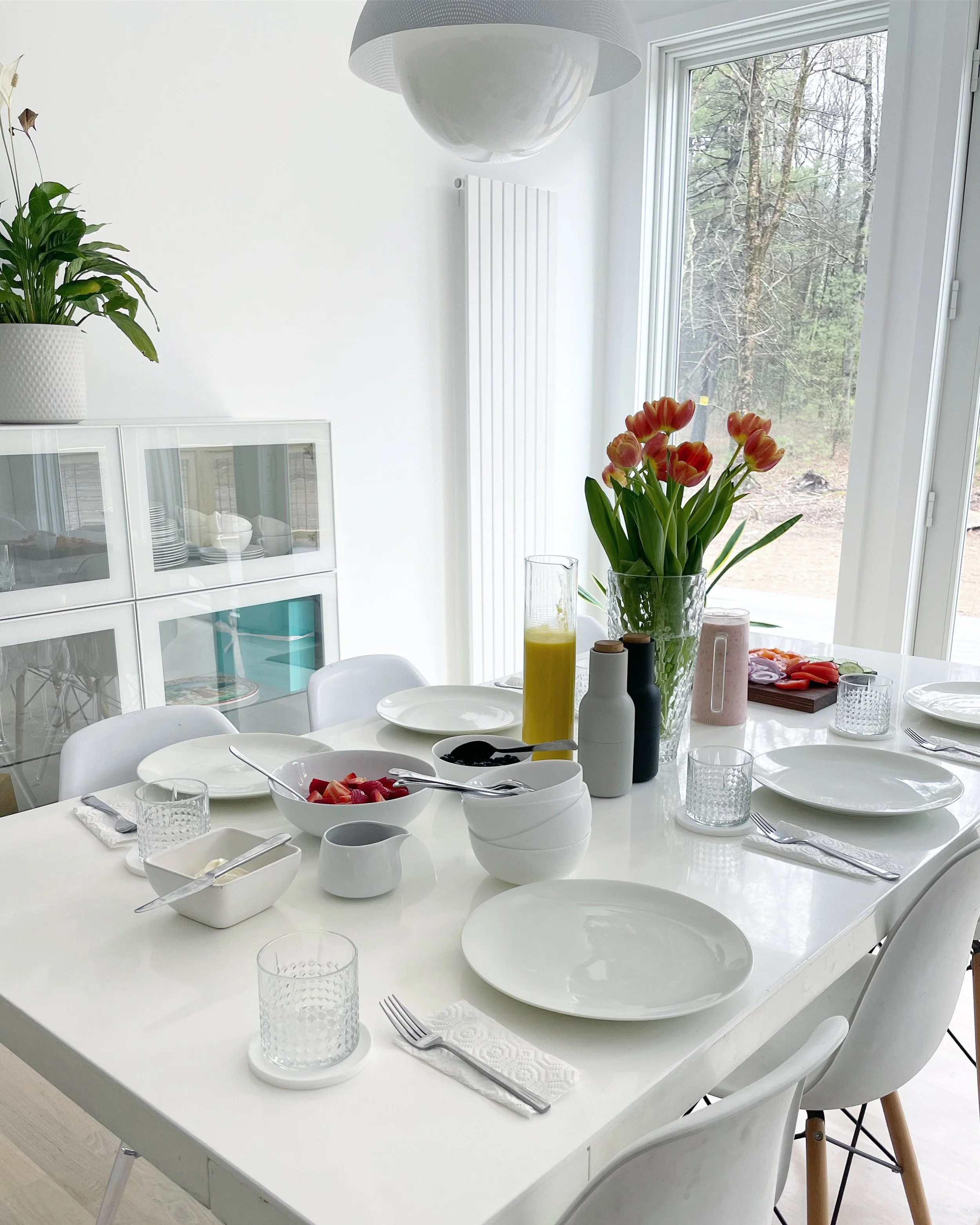 A bright, white dining room with a table set for breakfast, featuring white plates, bowls, glasses, utensils, a vase of pink tulips, a tall glass of orange juice, and condiments, near large windows overlooking a wooded backyard.