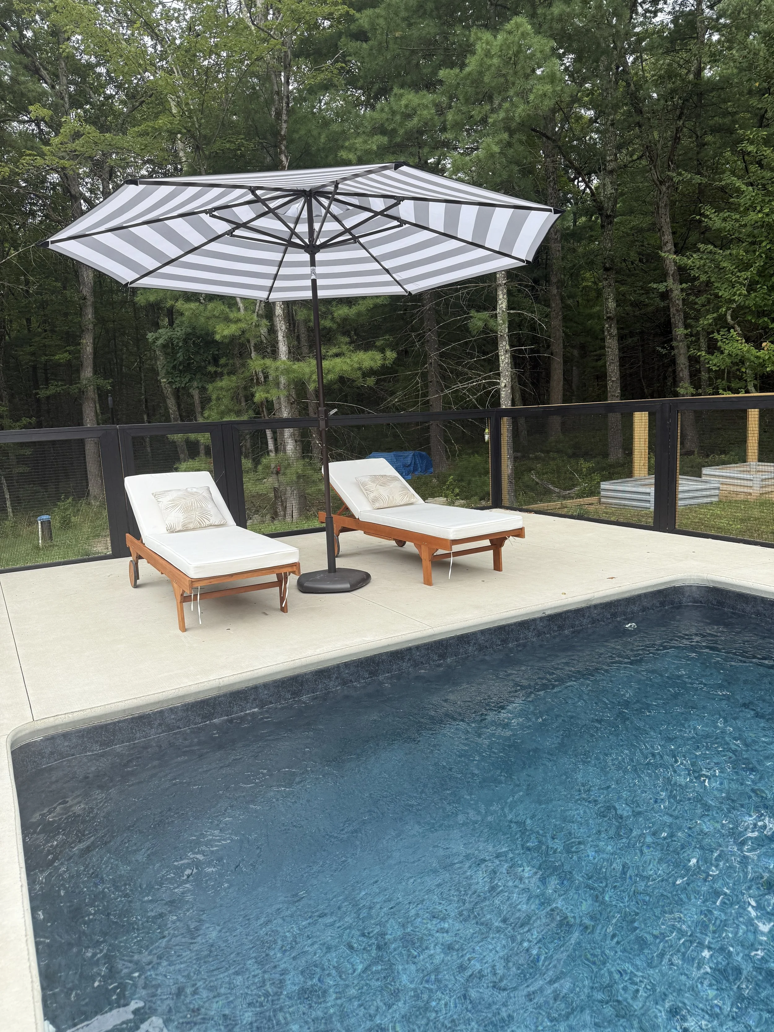 Swimming pool with two lounge chairs, an umbrella, and a wooded background.