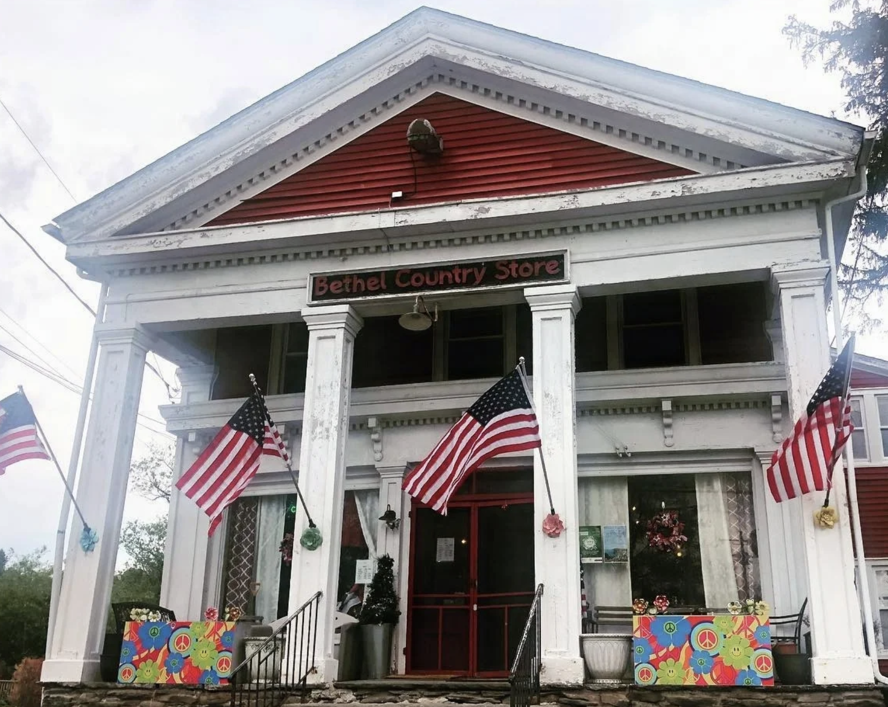 A two-story white building labeled 'Bethel Country Store' with American flags, colorful peace sign signs, and decorated with pink and green flowers outside.