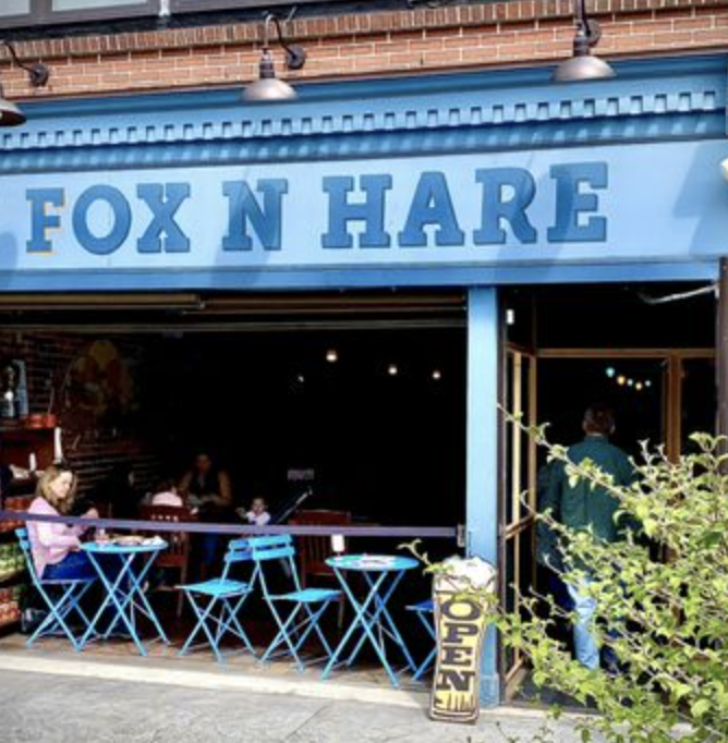 Exterior of a restaurant named Fox N Hare with an open entrance, blue outdoor tables and chairs, and a person walking inside, with an 'Open' sign outside.