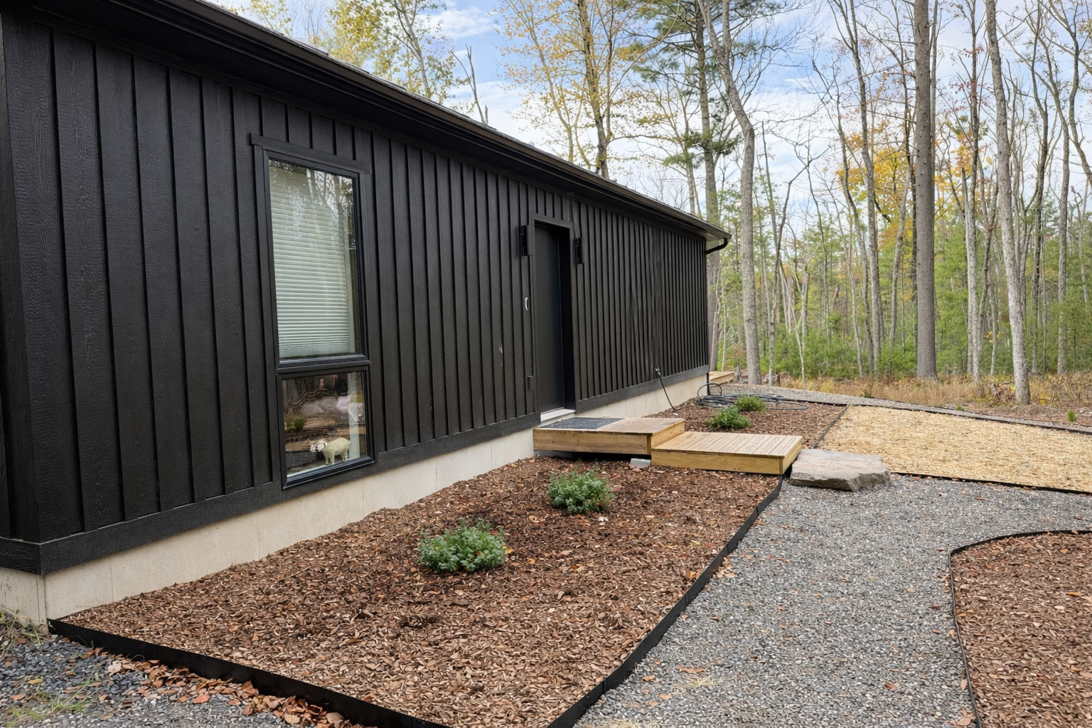 Backyard of a modern black house with a gravel pathway, small plants, and a wooded area with trees in the background.
