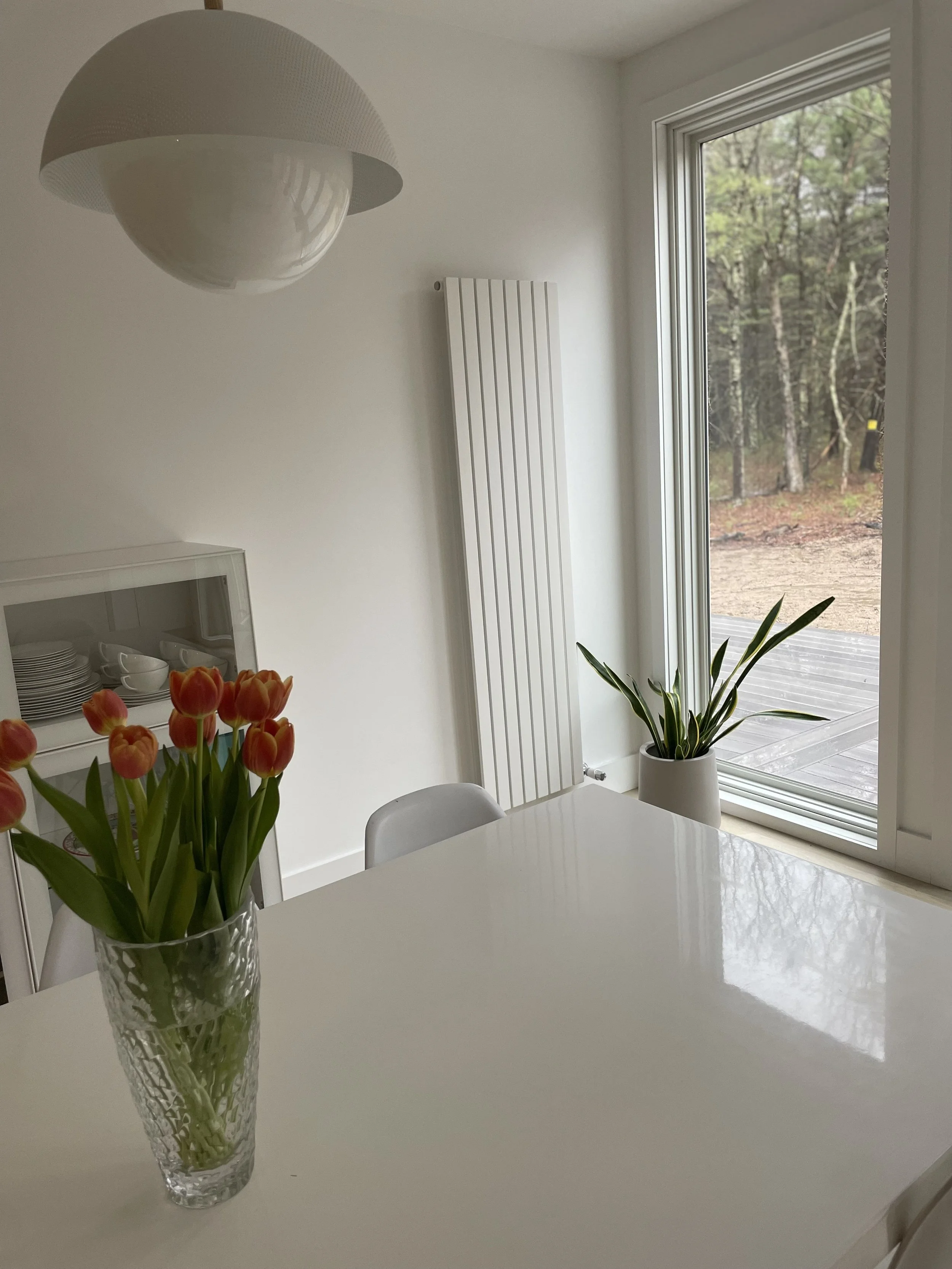 A dining area with a white table, a vase of pink tulips, a potted green plant near a large window, and a modern white hanging light fixture in a bright room with a view of a wooded outdoor area.