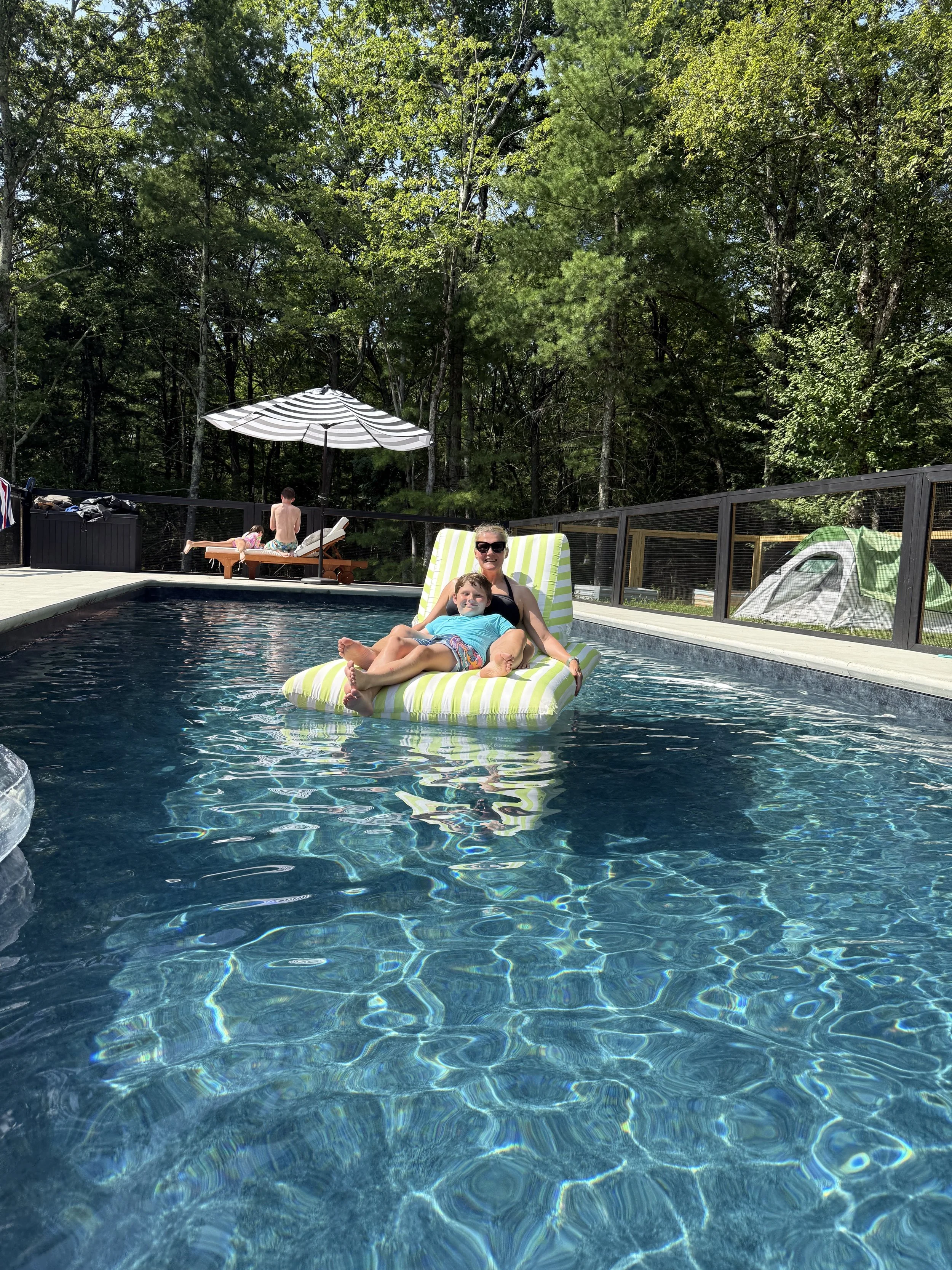 A woman and a young boy relax on an inflatable float in a backyard swimming pool, surrounded by trees. In the background, a child and a man are lounging on chairs under a large umbrella, and a tent is visible behind a fence.