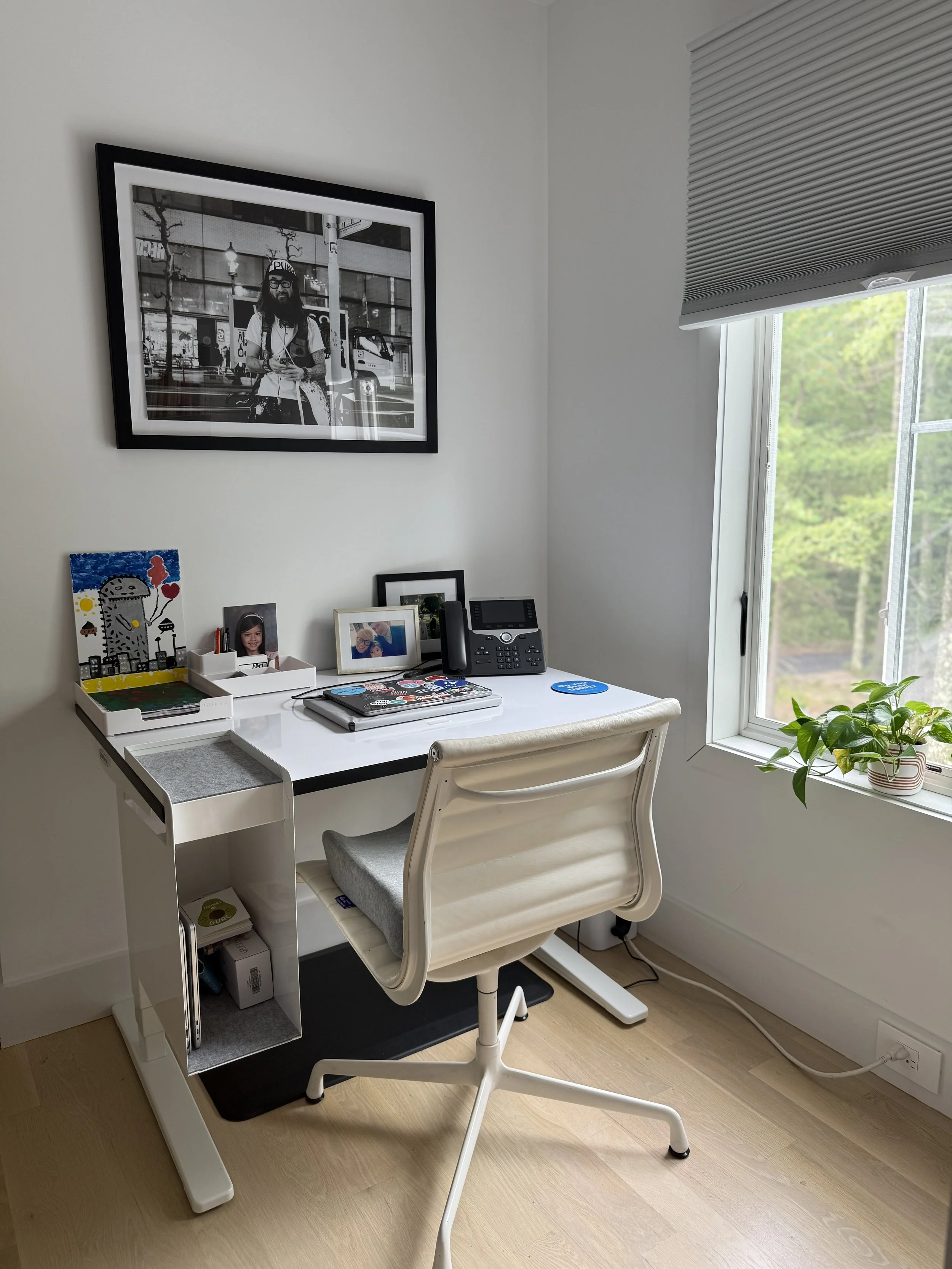 An organized home office with a white desk, beige office chair, framed photographs, a landline phone, and a potted plant on the windowsill. A large black-and-white framed photo hangs on the wall.