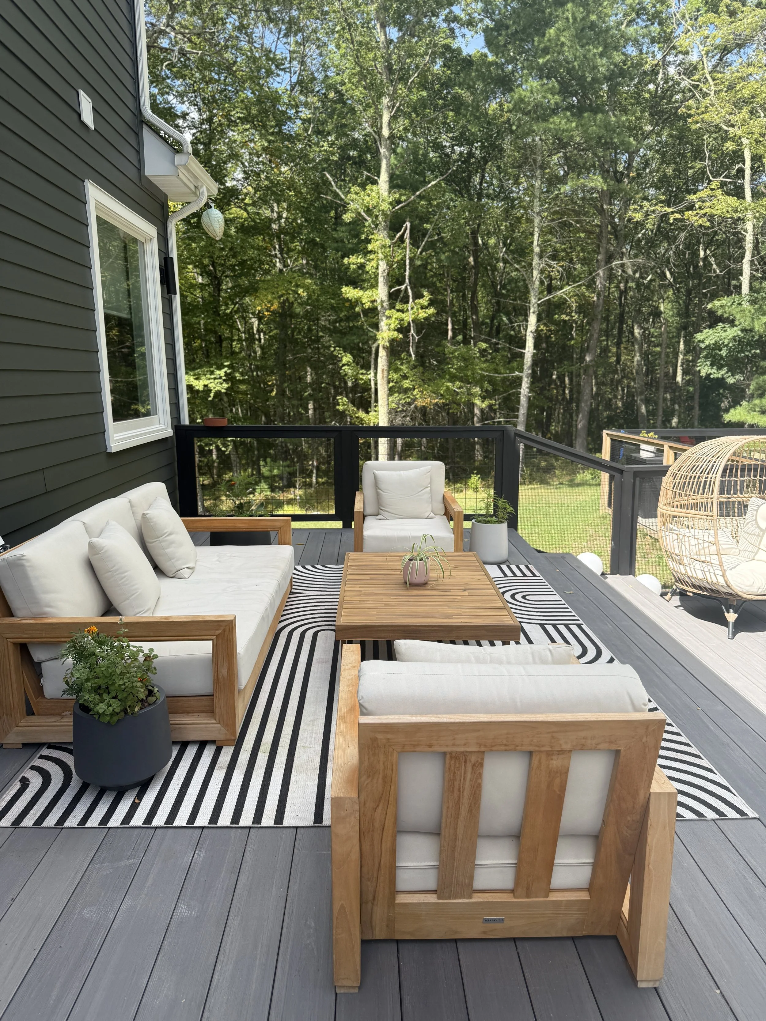 Outdoor deck with wooden furniture, including a sofa, two chairs, a coffee table, and a hanging egg chair; black and white striped rugs; potted plants; surrounded by green trees.