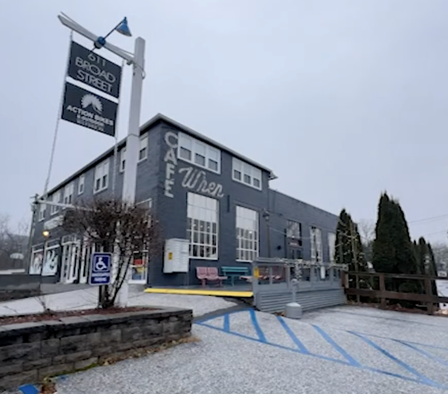 A gray building with large windows, featuring signs for 'Old Road Street' and 'Wilson.' A small seating area with pink and blue benches is outside. There is a handicap parking sign and blue markings on the parking lot in front.