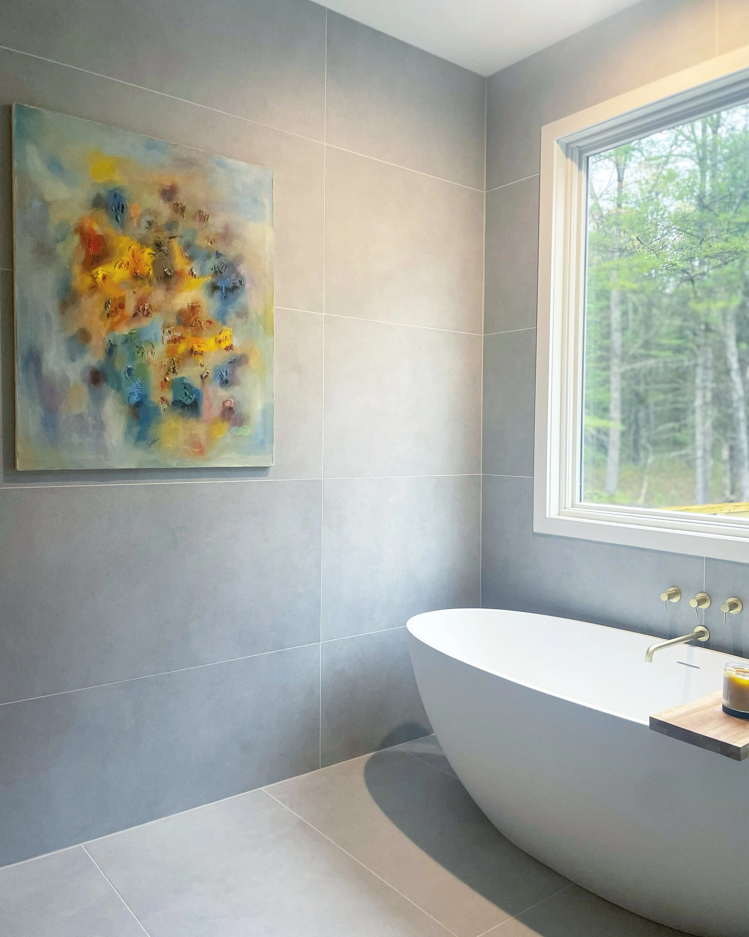 Modern bathroom with a white bathtub, gray tiled walls and floor, a large window showing trees outside, and colorful abstract art on the wall.