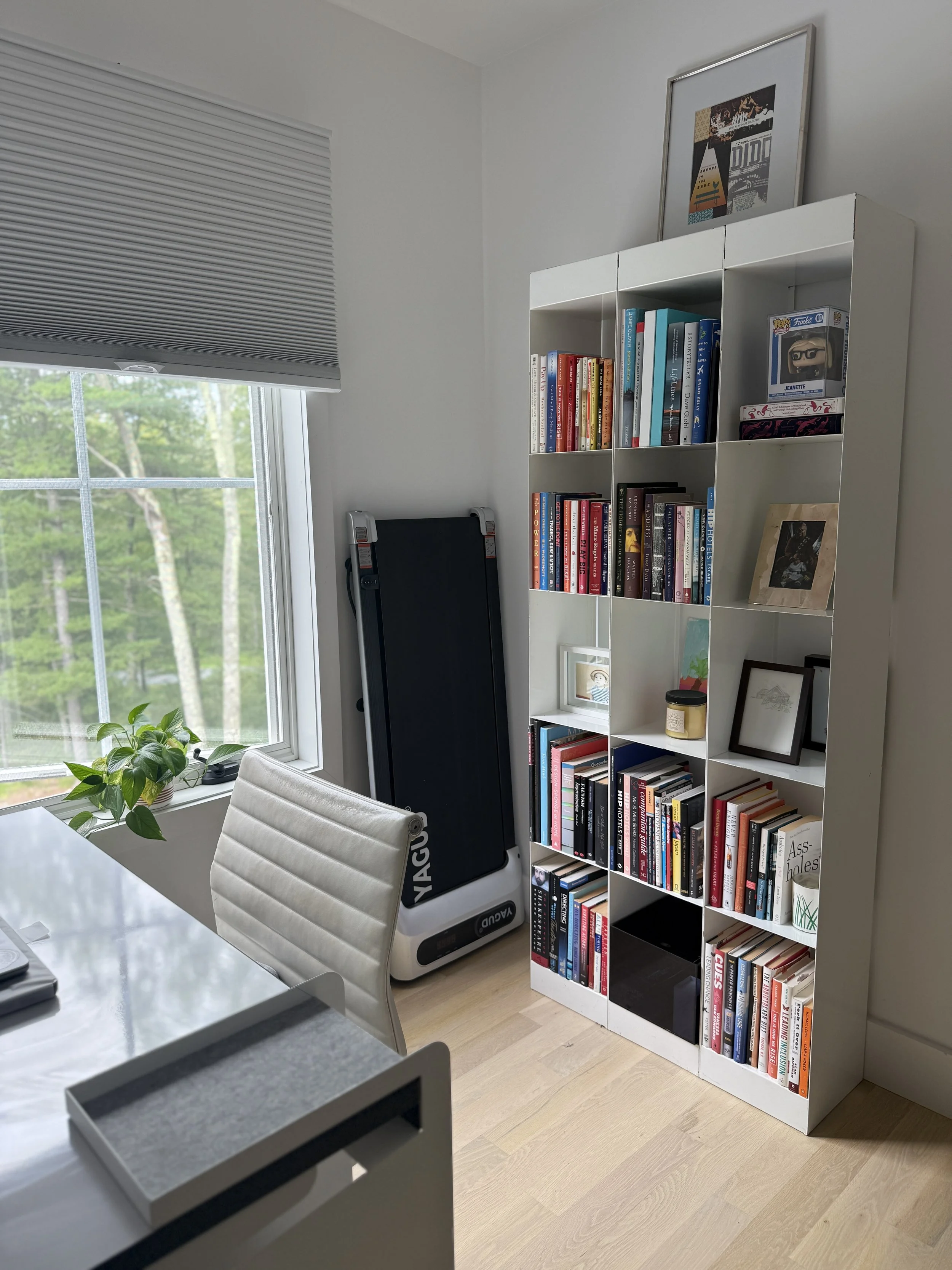 A white bookshelf filled with books, a framed picture, and decorative items in a room with a large window and a white office chair.
