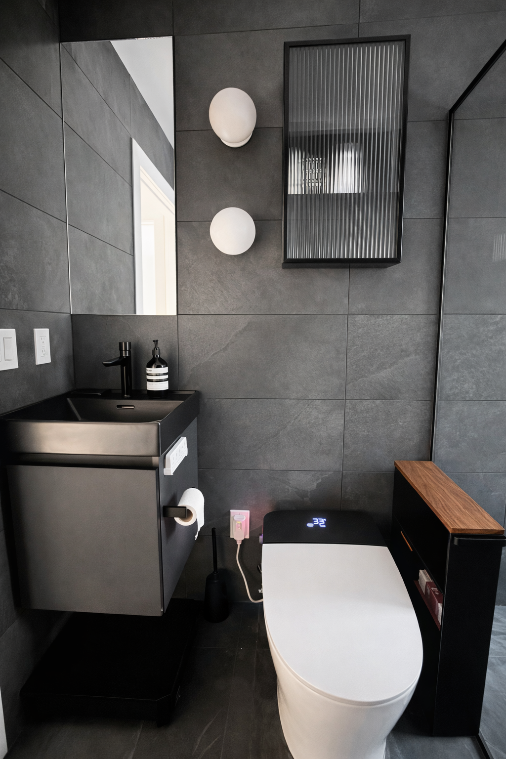Modern bathroom with dark gray tiled walls, a sleek black sink, a mirror, wall-mounted light fixtures, a digital bidet toilet, and a wooden shelf.
