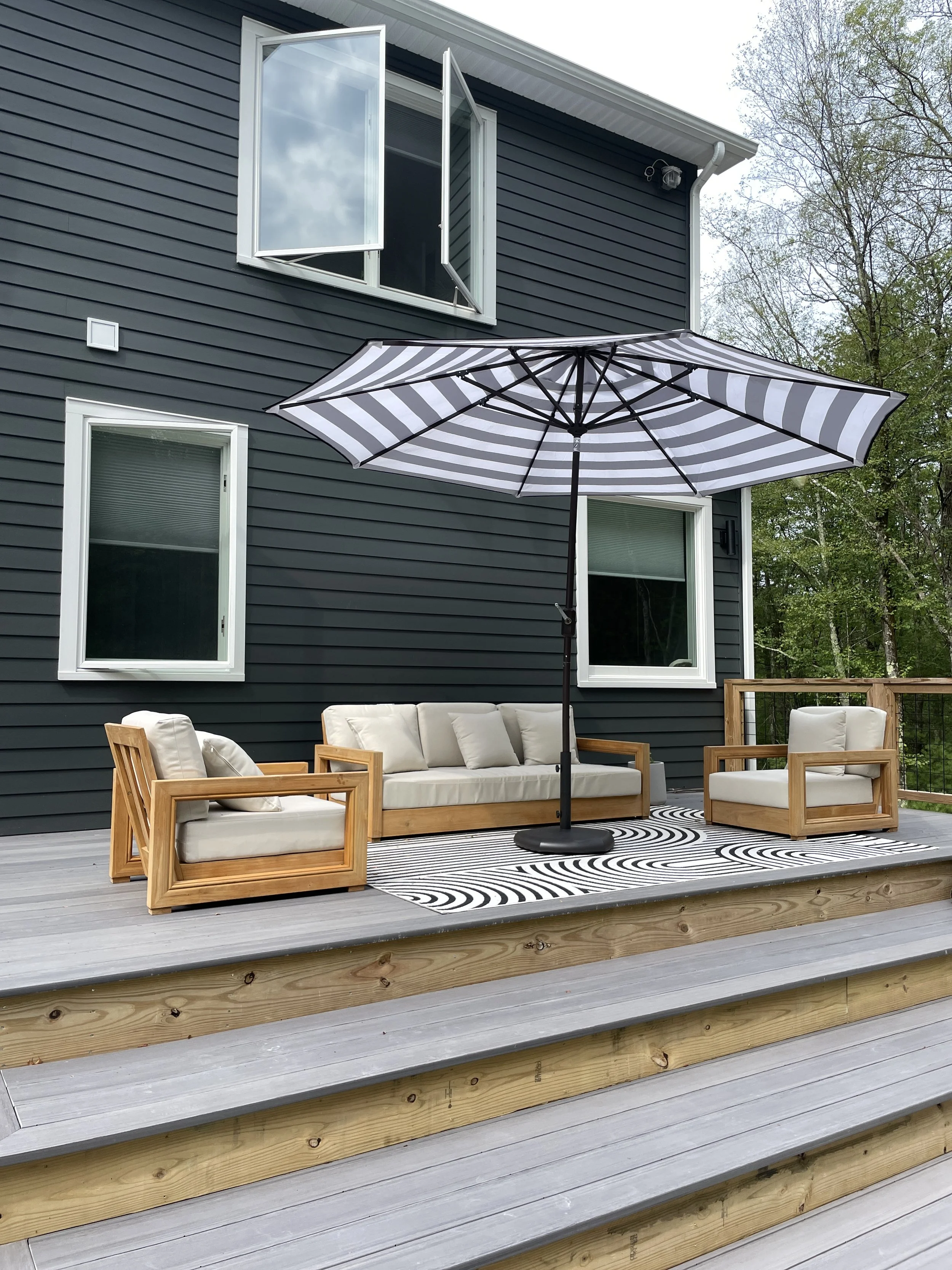 Outdoor deck with wooden furniture, cushions, black and white striped umbrella, and a patterned outdoor rug, adjacent to a dark blue house with open windows and a wooded background.
