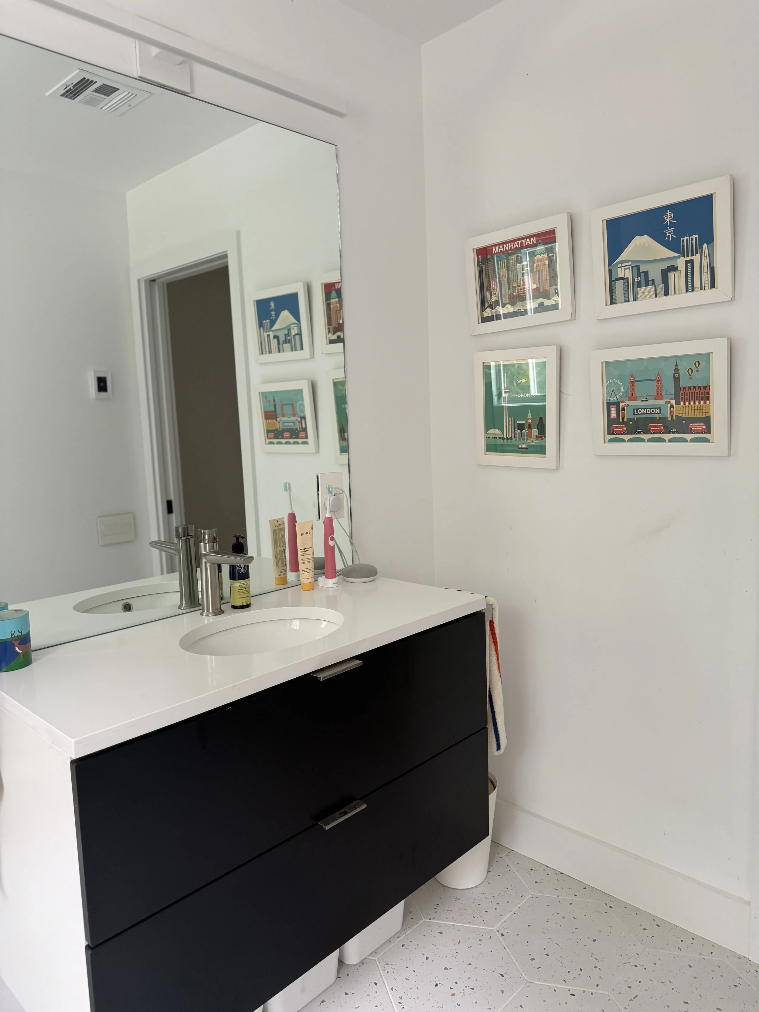Bathroom with a large mirror, black vanity with a white countertop, bottles of toiletries, and framed cityscape prints on the wall.