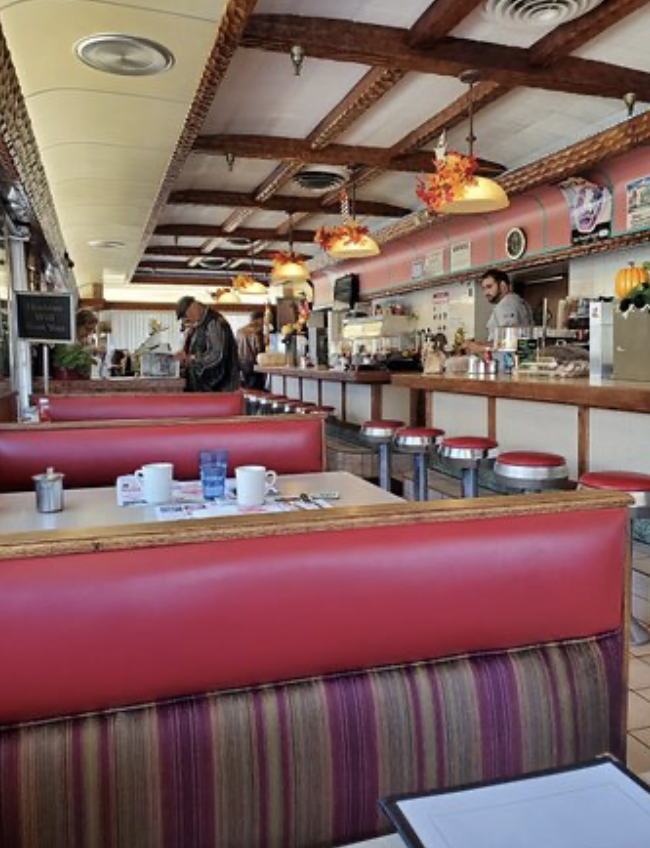 Interior of a retro-style diner with red booths, a long counter with bar stools, pendant lights, and customers ordering food.