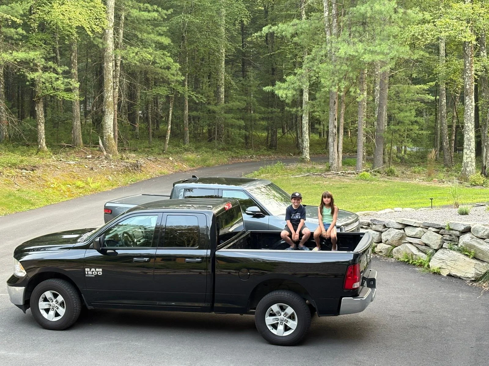Two children sitting in the bed of a black RAM 1500 pickup truck parked on a paved driveway, with a wooded forest background.