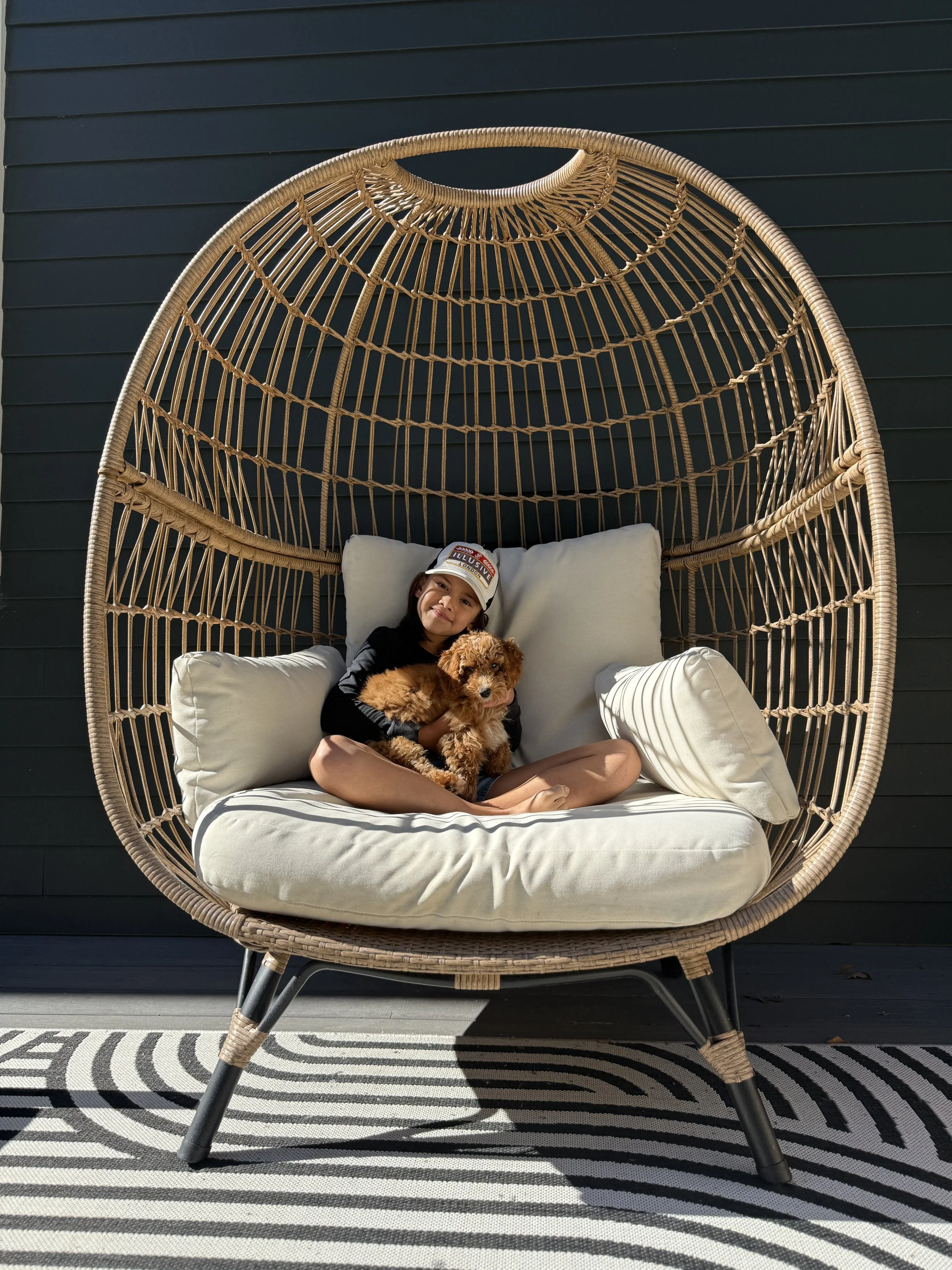 A girl sitting on a cushioned outdoor wicker chair holding a small brown puppy, with a dark blue exterior wall behind them and a black and white striped outdoor rug underneath.